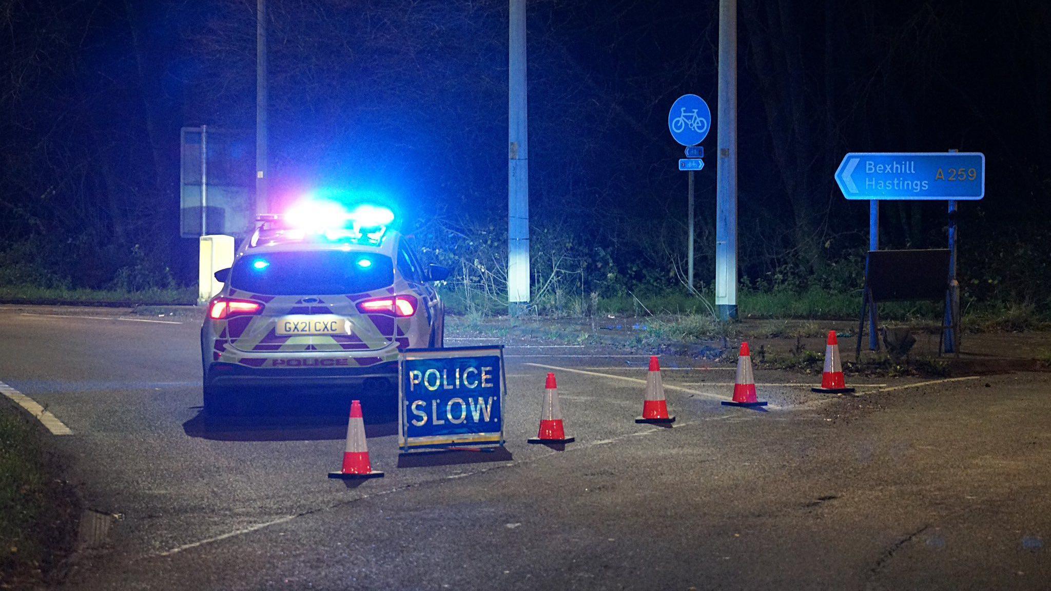 A police car parked with its lights shining blue, a sign which says POLICE SLOW next to some orange and white traffic cones blocking a road and a sign pointing towards Bexhill and Hastings