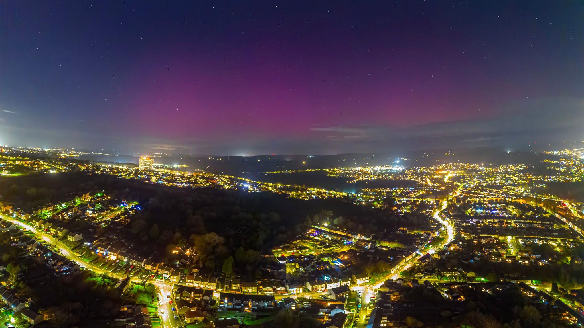 A large patch of pink sky seen in darker skies above brightly lit city, with street lights and traffic lighting up the roads seen from above