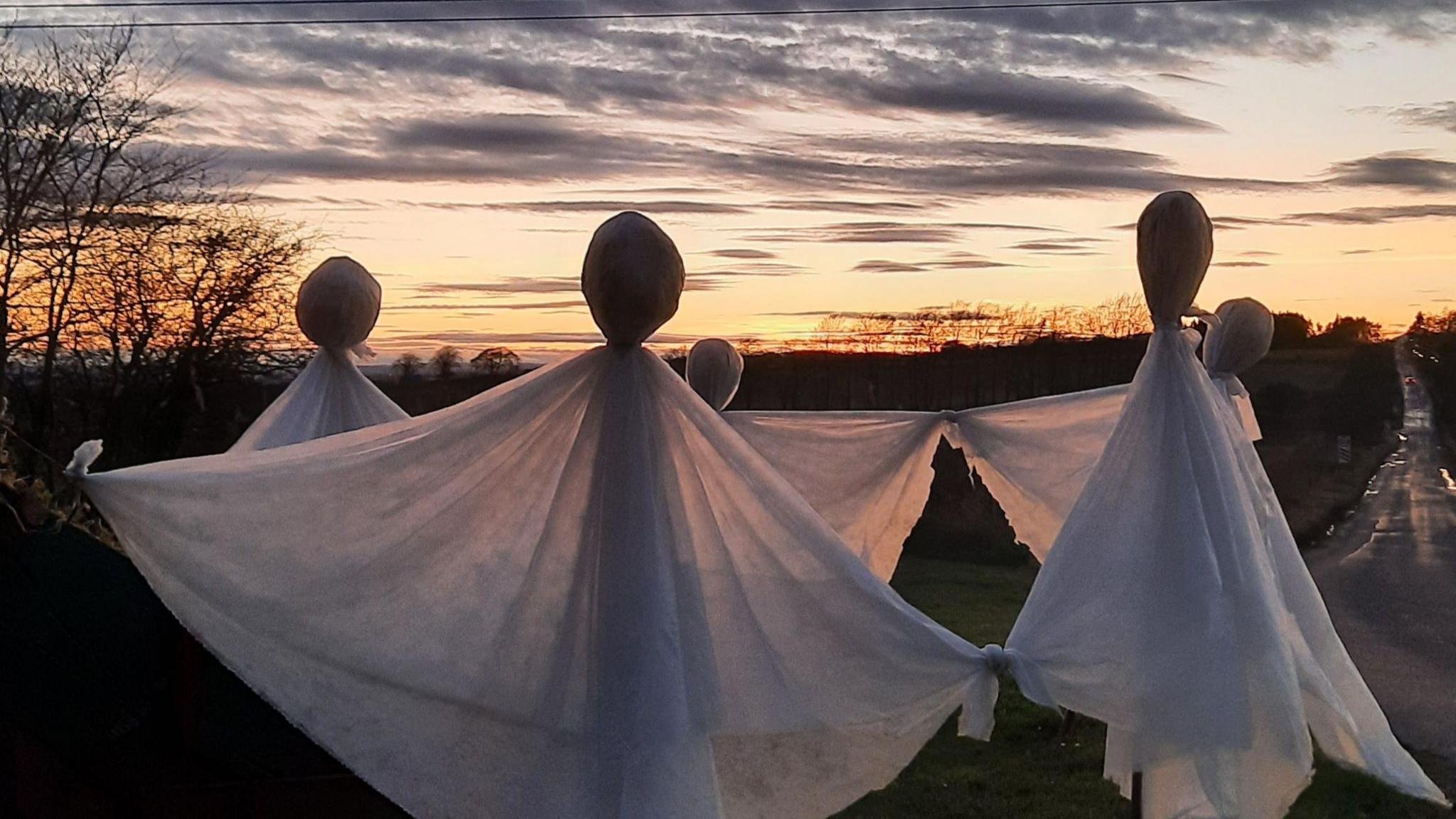 A sculpture of three ghosts at dusk. There are three sticks emerging from the ground half way up the picture. They are each covered with a white sheet, which is pulled out at either side. At the top, they are tied in to a circle to represent the head. Above them are dark clouds with orange light in the distance.