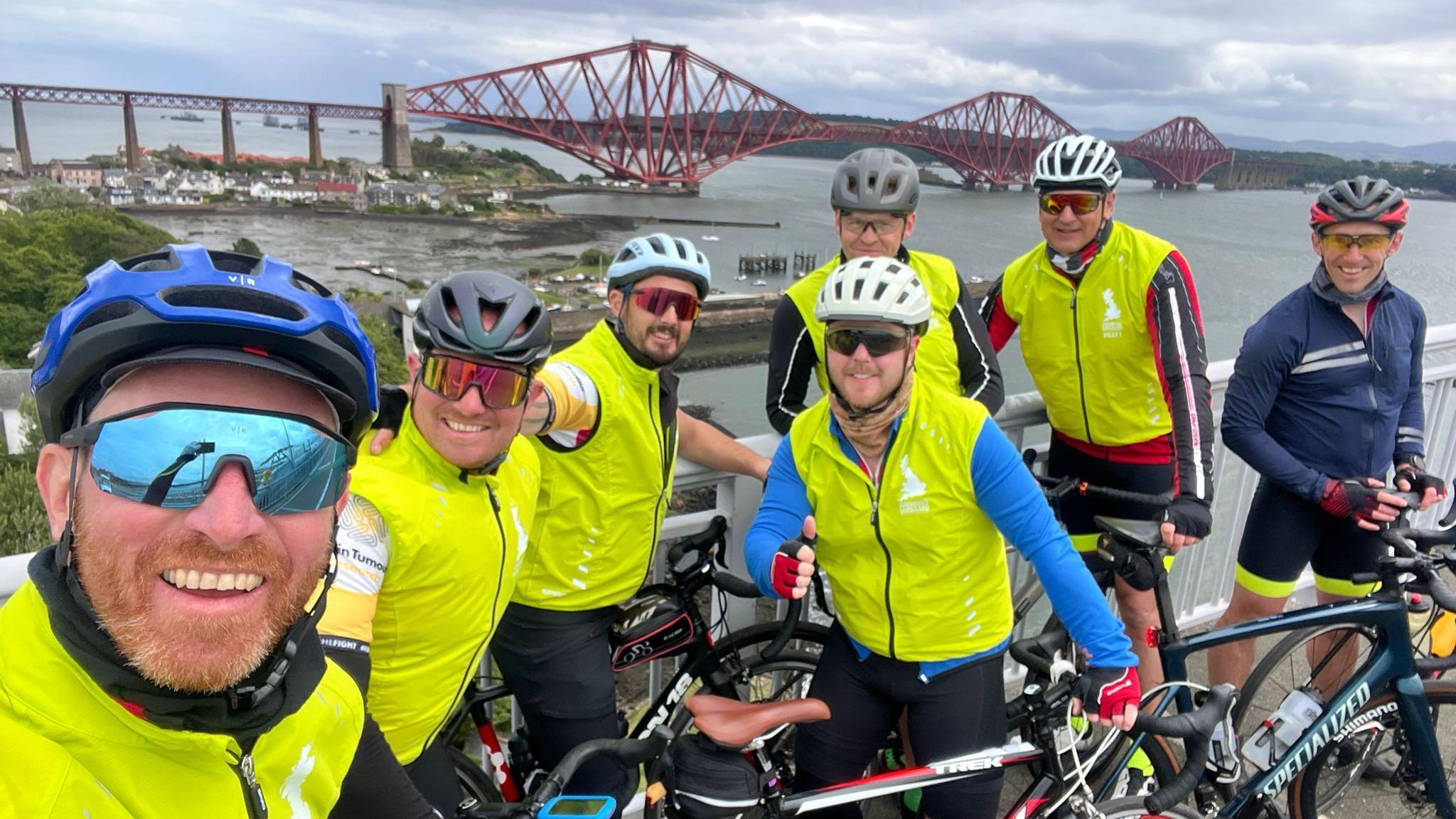 A group of seven men wearing high-visibility jackets smiling at the camera with their bikes, with the Forth Bridge in the background.