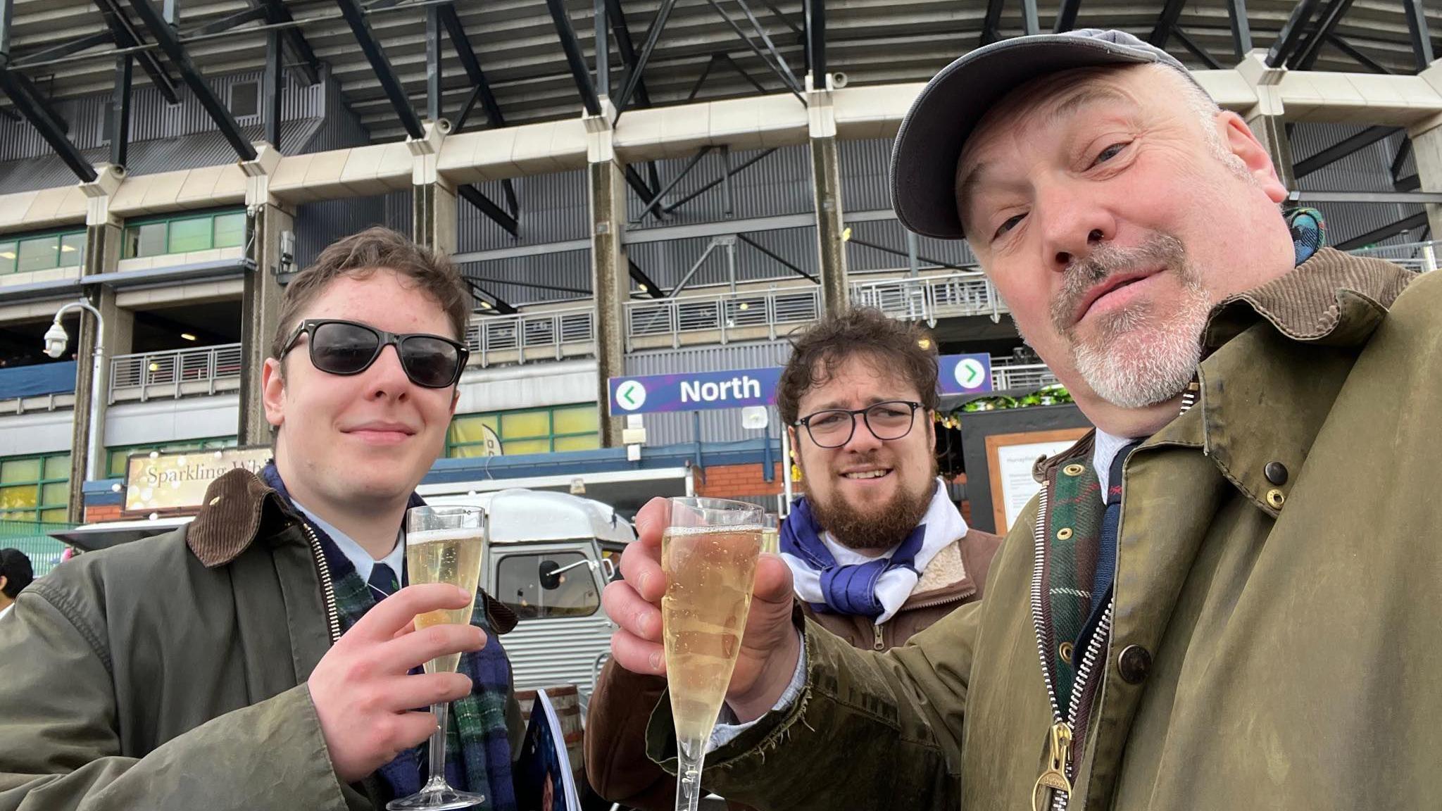 A young man wears sunglasses to the left, holding a drink next to another young man wearing a Scotland scarf and then to the right an older man wearing a cap, holding a drink