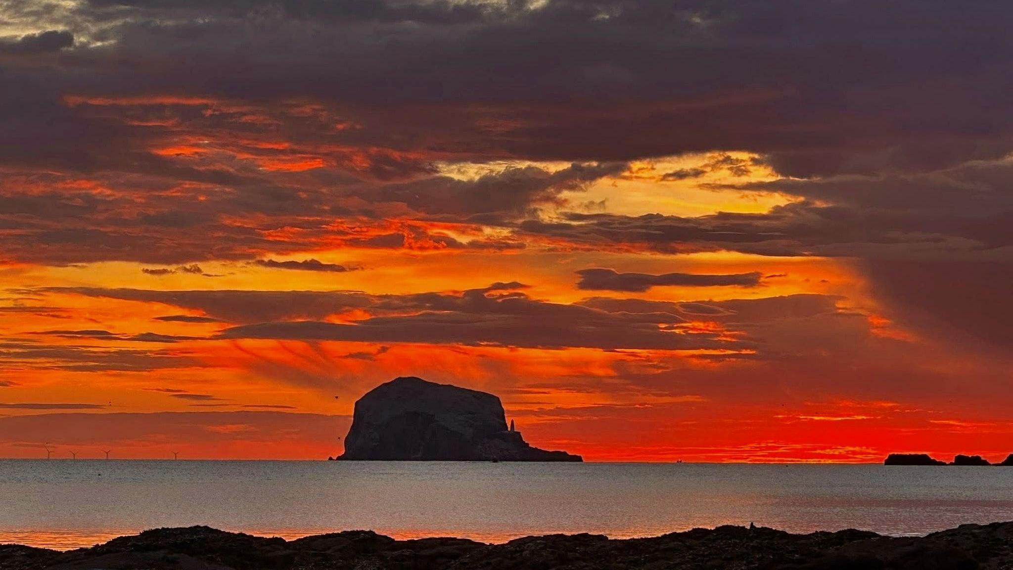 The Bass Rock forms a dark silhouette against an orange sky that looks like its on fire. The sea is in the foreground.
