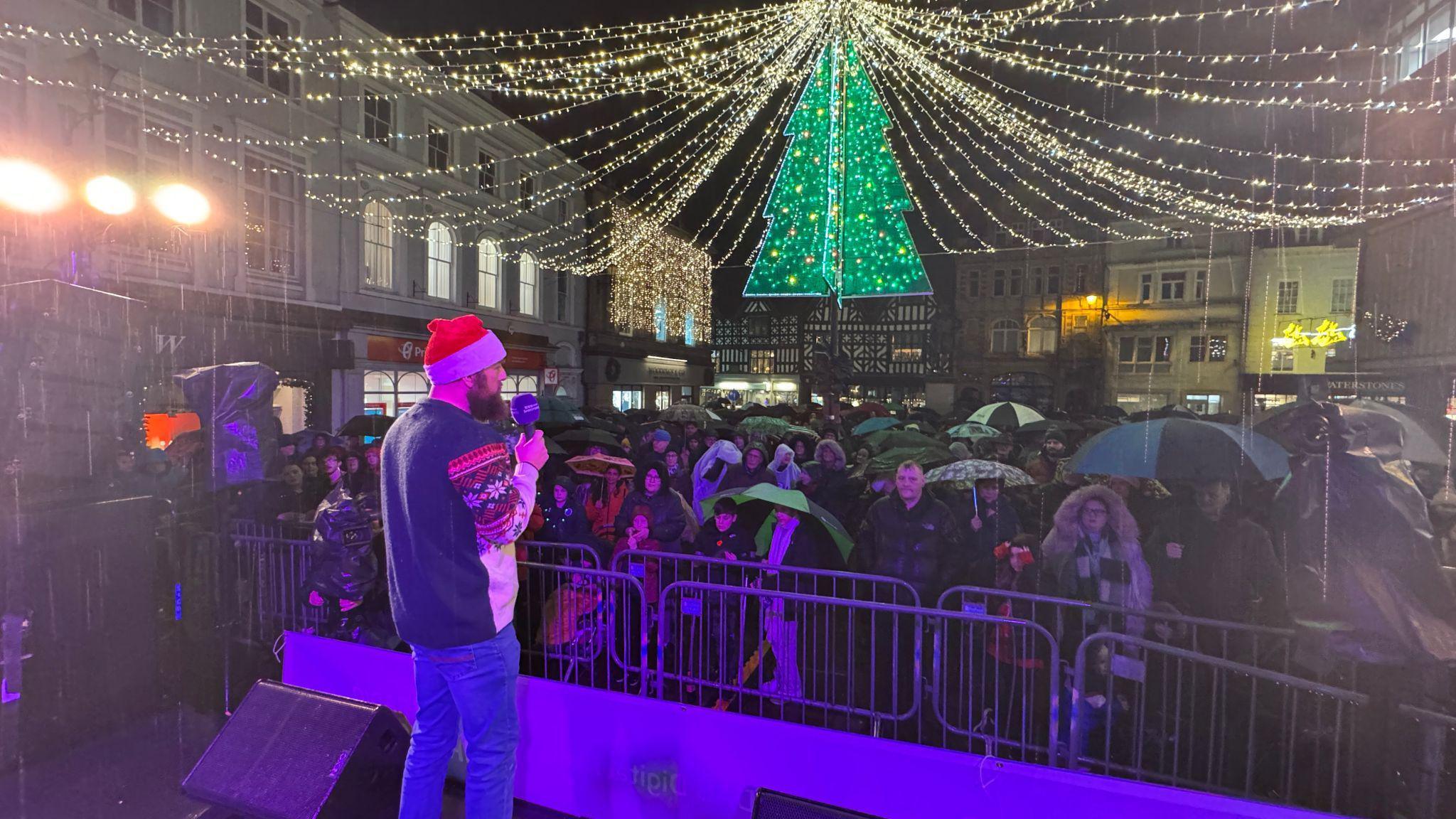 A man in a Santa hat and a beard, wearing a Christmas jumper and jeans, holds a microphone in front of metal railings. Behind them, a crowd of people hold umbrellas with Christmas lights on chains running overhead, peaking around a lit-up sculpture of a Christmas tree.