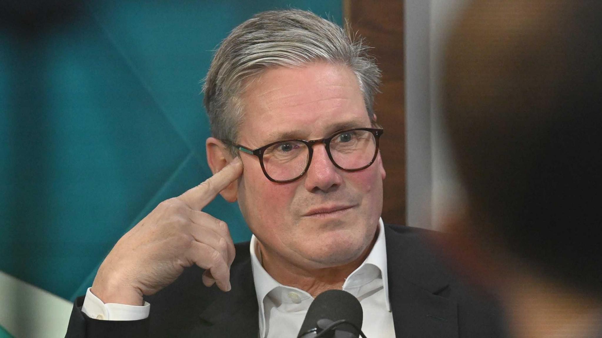 Keir Starmer sitting in front of a microphone with his fingers pressed against the right side of his temple, wearing an open-necked shirt and dark suit jacket