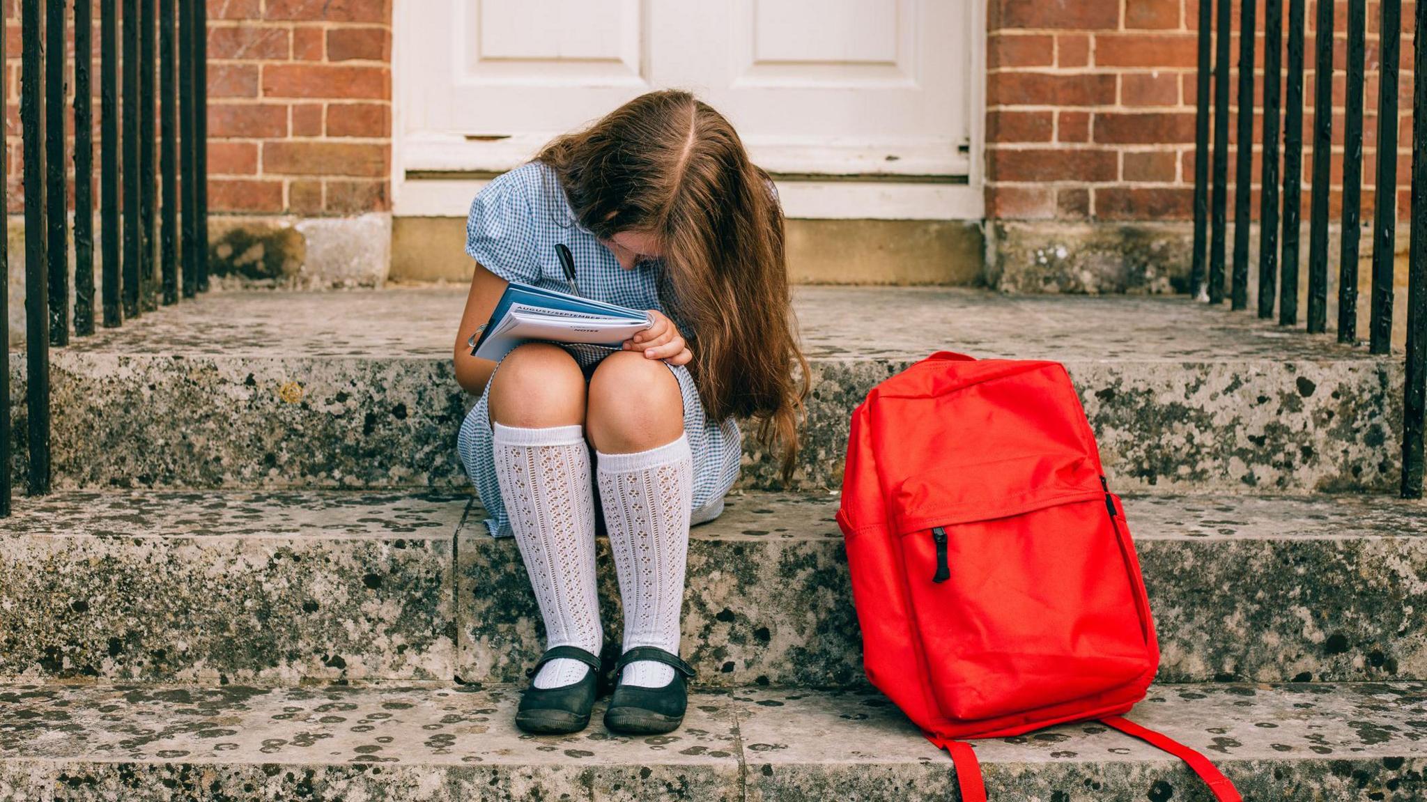 a girl in a blue and white school uniform doing some schoolwork sitting on a step, her bright red rucksack is next to her