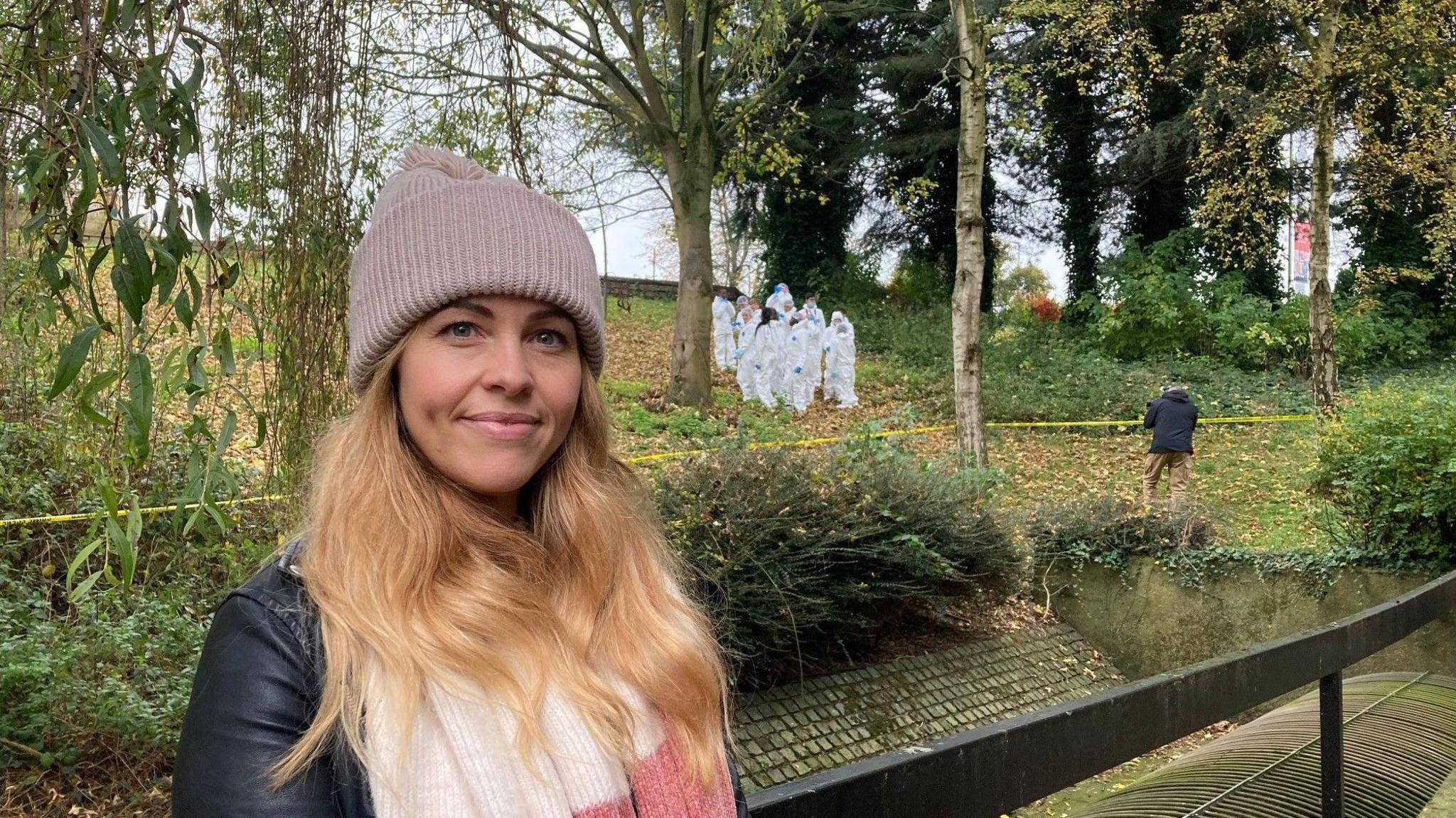 A blond-haired woman wearing a light pink woollen hat and a black leather jacket and cream and pink scarf in the foreground. Pupils in white Scenes of Crime suits walk up a grassy hill in the background.