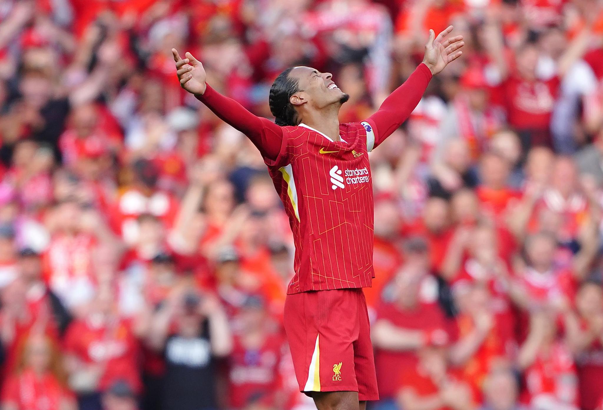 Virgil van Dijk wears red football kit and holds his arms to the air smiling, with a crowd in red blurred out behind him.