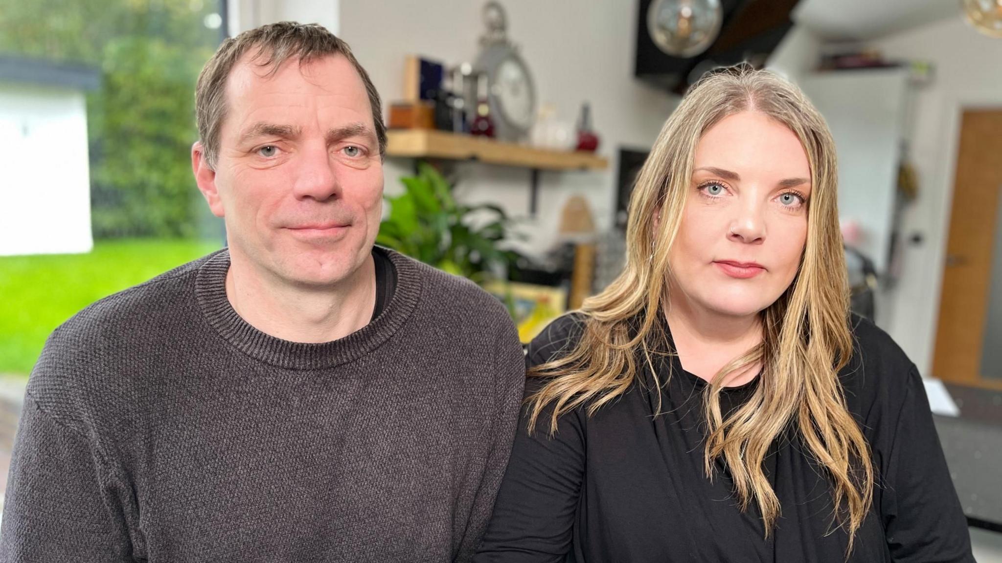 Dave and Chloe Longster look at the camera in a house. Mr Longster, on the left of the picture, has short brown hair and is wearing a brown wool jumper. Mrs Longster has long, blonde, wavy hair and is wearing a black long-sleeved top.