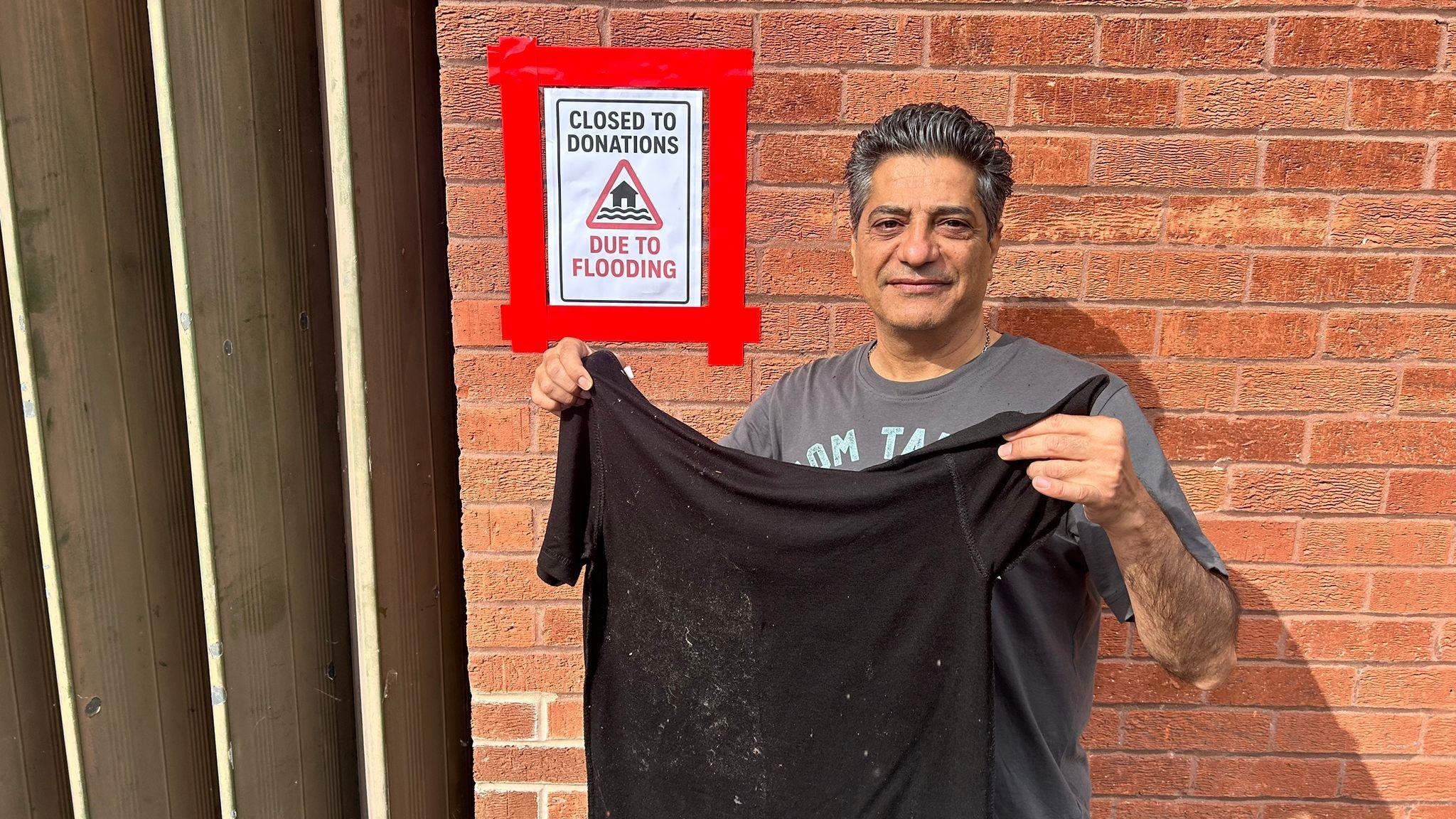 A man stands in front of a wall holding a black t-shirt which has marks on it. There is a sign behind him which reads 'Closed to donations: Due to flooding".
