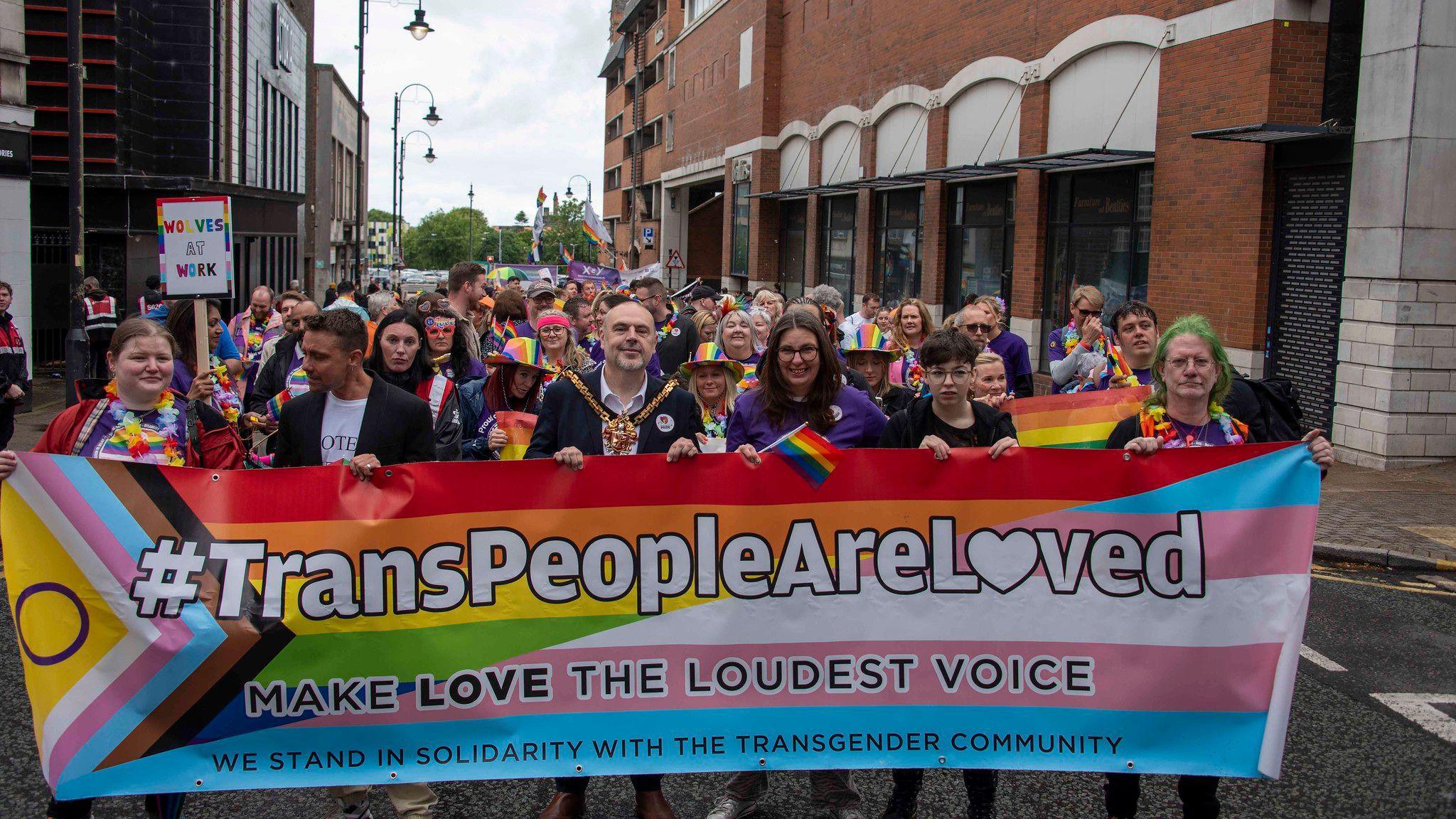 People walking towards the camera through a city centre street as part of a parade. Many are wearing rainbow colours and purple shirts. One man at the front is wearing a gold mayoral chain. Six people at the front are holding a large multi-coloured banner which says "#TransPeopleAreLoved, make love the loudest voice, we stand in solidarity with the transgender community".