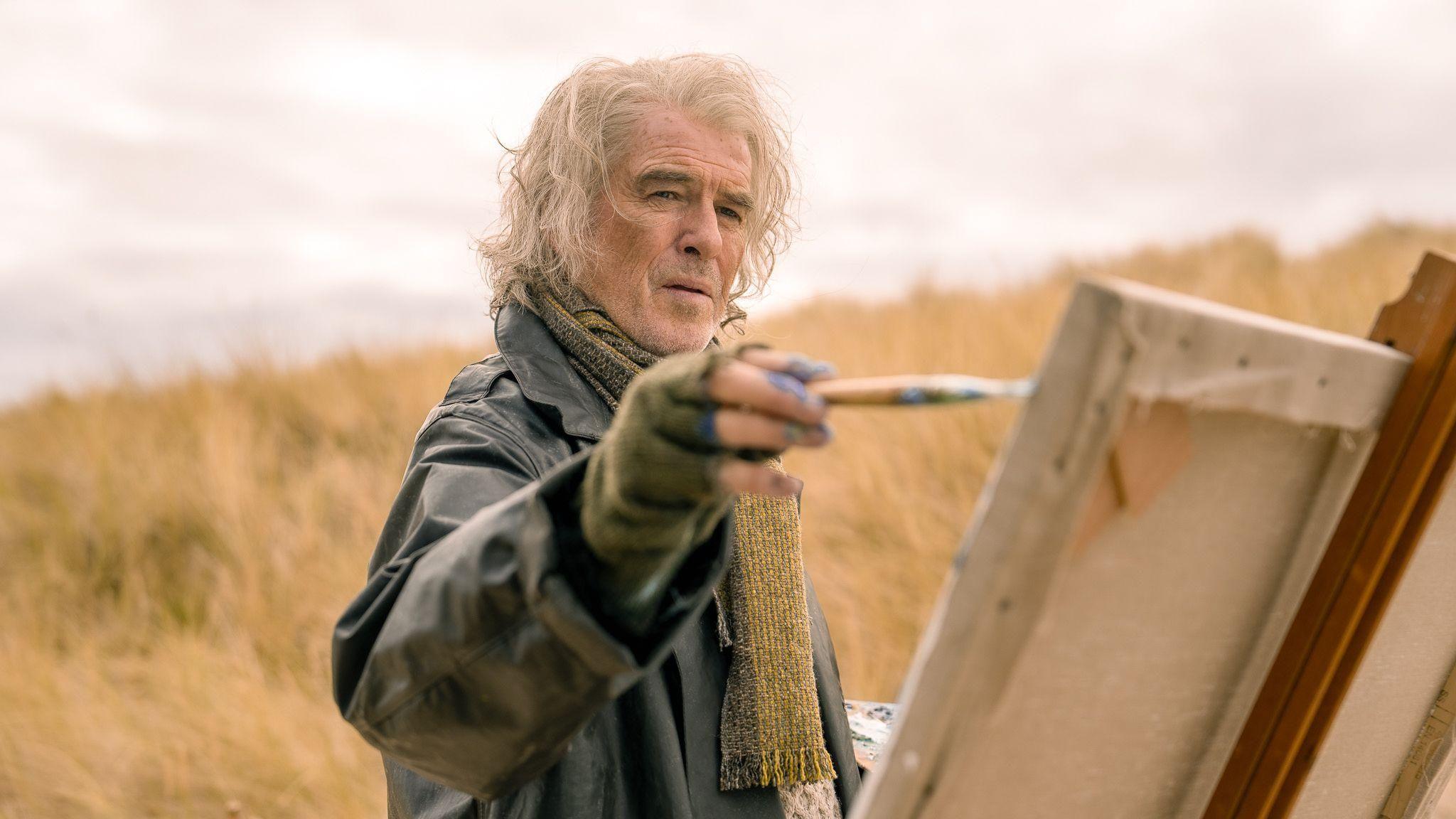 A still of Pierce Brosnan in the film. He is wearing a  black leather jacket and a green scarf. His hair is white and worn long. He is using a paint brush on a canvas supported by an easel. In the background is long yellow grass that looks like it is a sand dune.