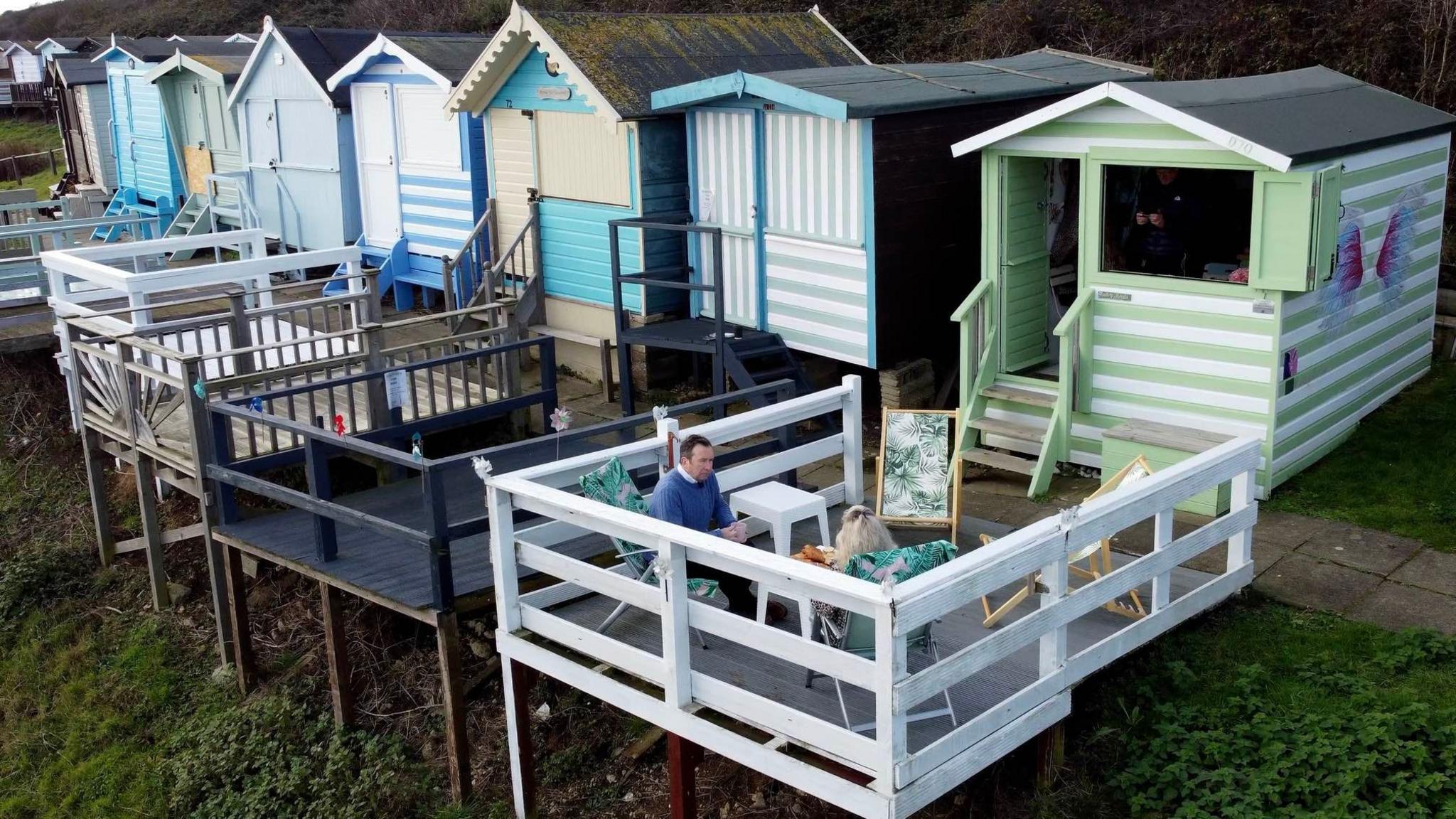 A picture taken by a drone of a man and a woman sat on a balcony in front of a green and white-striped beach hut. There are several other colourful huts in the same row.