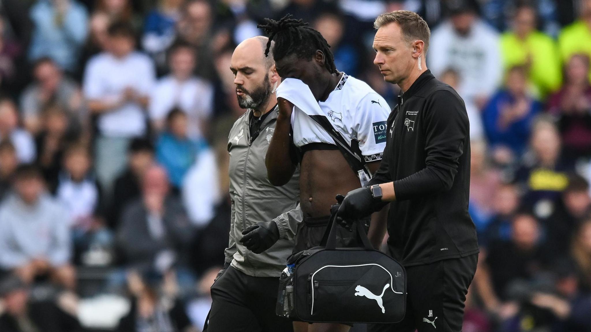 Derby County's David Ozoh coming off injured wiping his face with his shirt alongside two club medical officials.