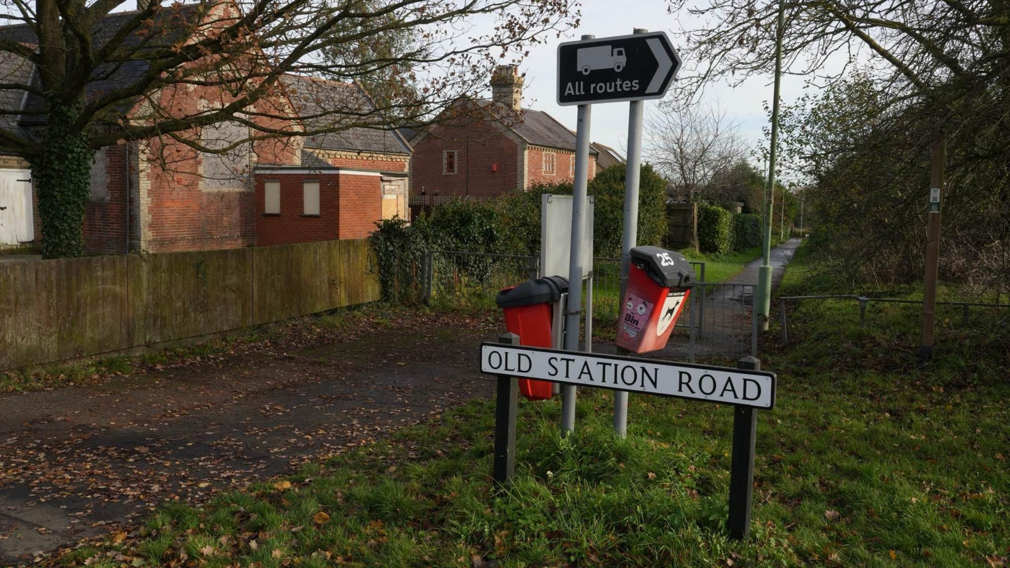 A concrete footpath running diagonally from left to right into the distance. There  are brown leaves on the path and houses to the left of the footpath. There is a brown All Routes sign and two red dog poo bins. A white street sign in centre states Old Station Road in black letters.  