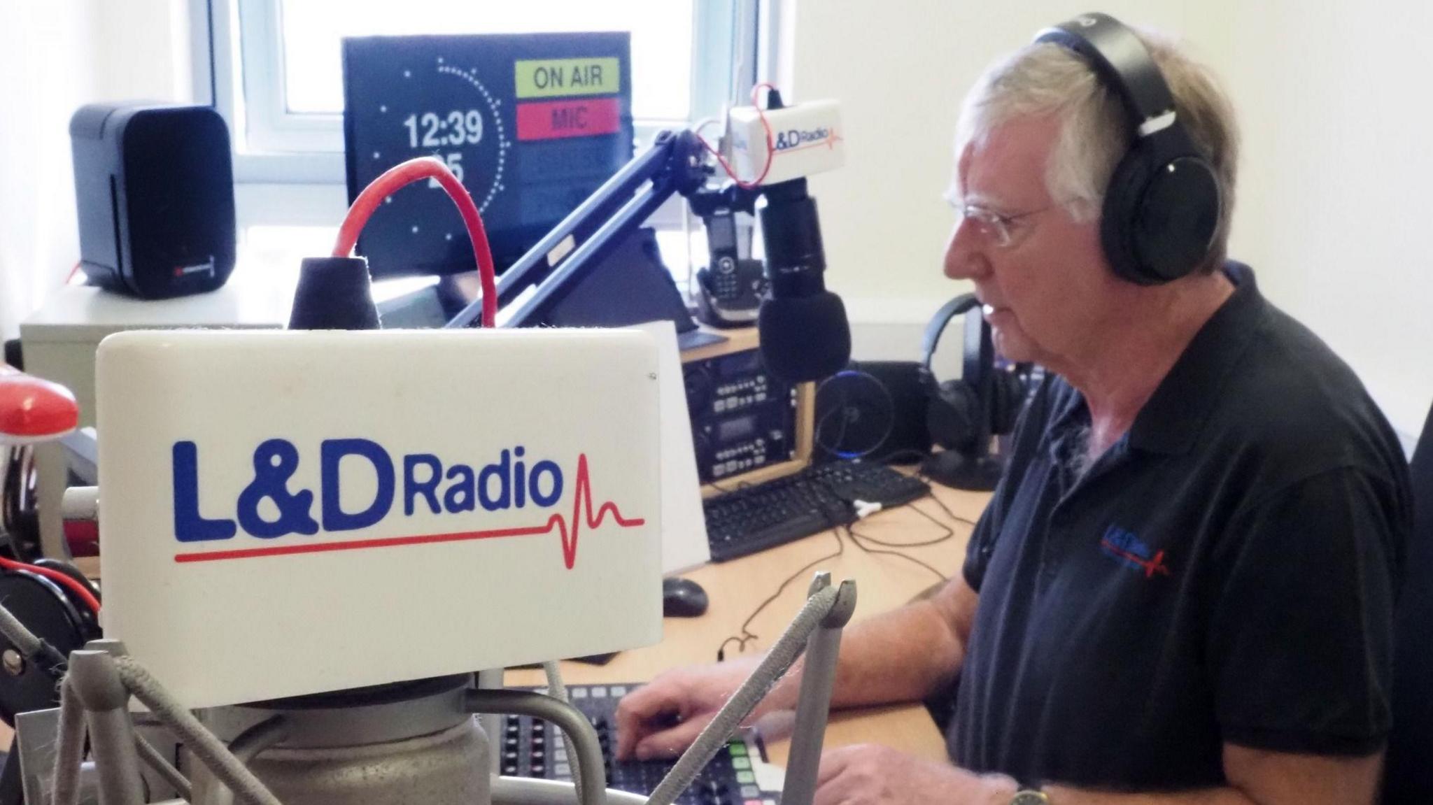 A man sits in a radio studio surrounded by speakers, a screen and a microphone. He is wearing headphones. There is L&D Radio branding