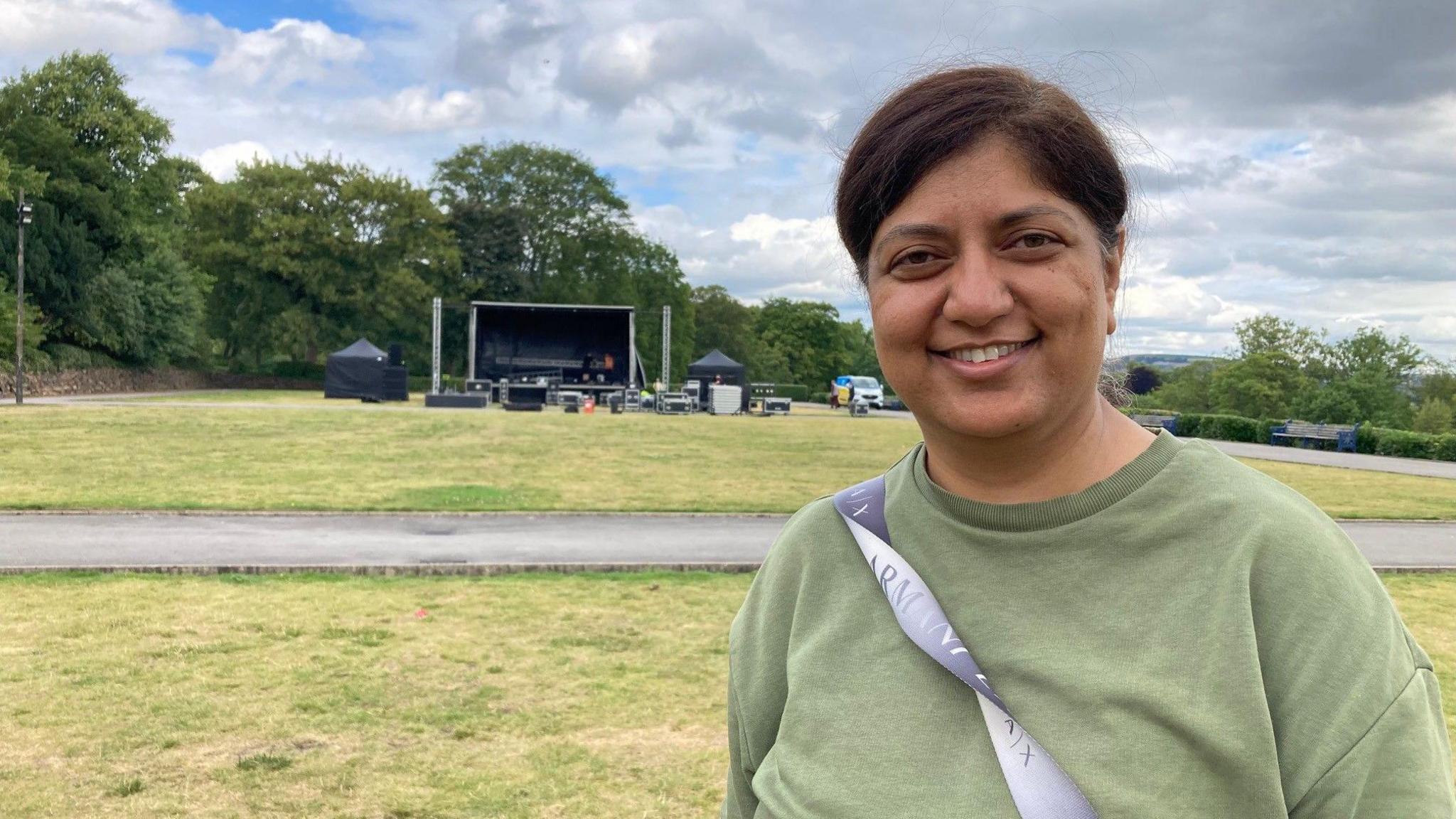 A woman with tied-back brown hair and wearing a green sweatshirt standing on some grass with an empty stage in the background.