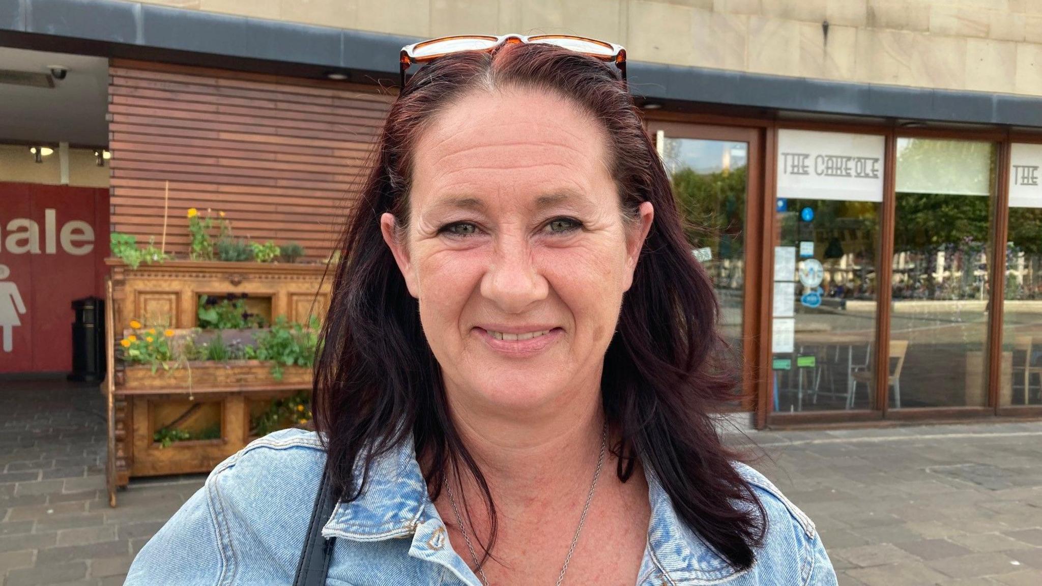 A white woman wearing a denim jacket stands outside a cafe. She has shoulder length brown hair and a pair of glasses perched on her head