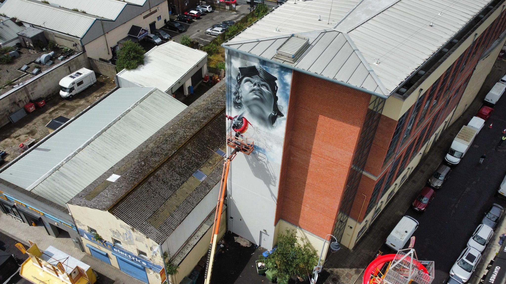 Artist jeks stands on a cherry picker while painting a mural of Amelia Earhart on the Foyle building in Derry, in the backgournd are the city's rooftops