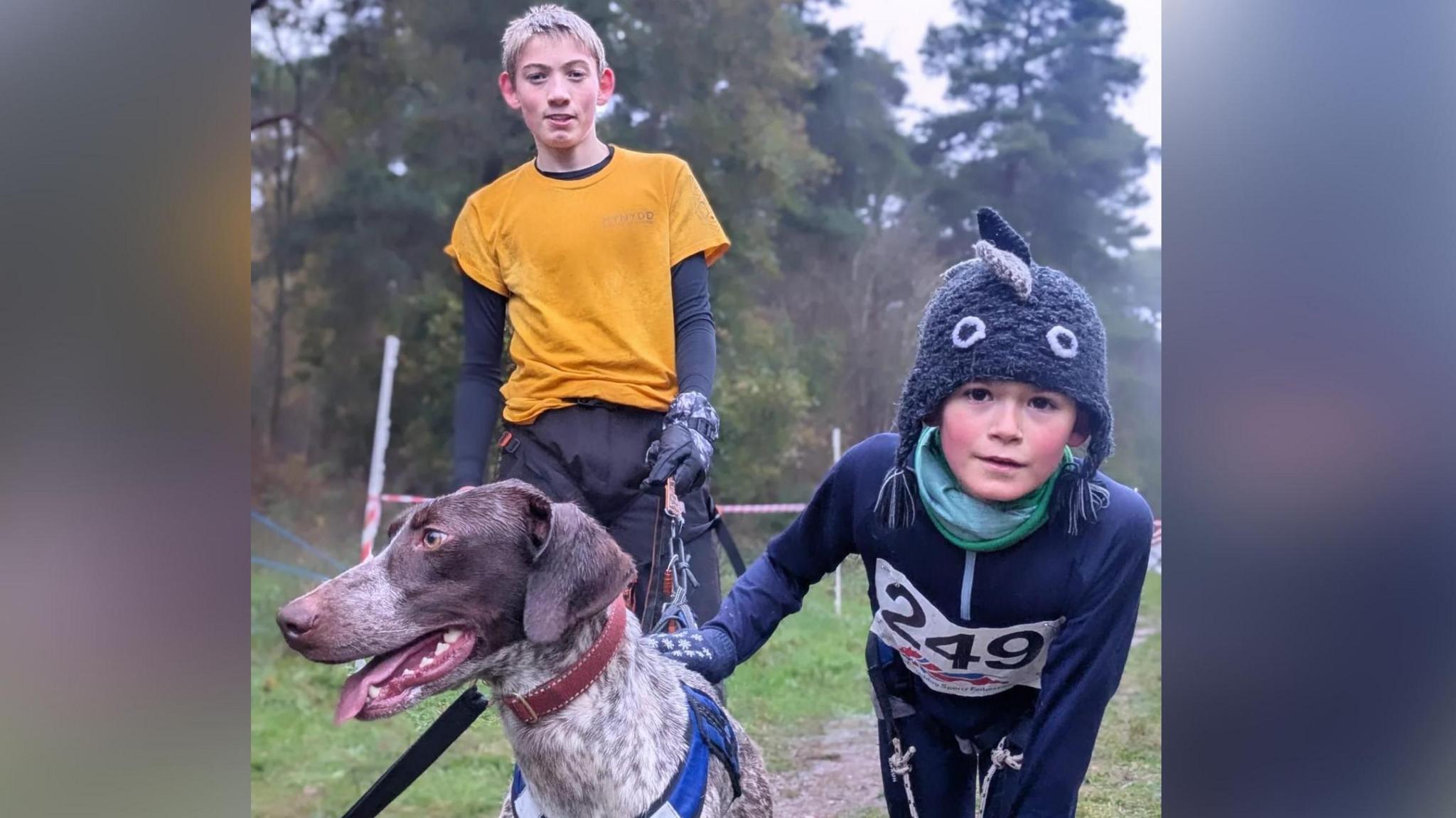 Llun o gi a ddau fachgen. Mae un bachgen yn sefyll tu ôl i'r ci mewn crys-t melyn a throwsus du. Nesaf i'r ci mae'r bachgen arall sy'n gwisgo het a siaced glas tywyll. Maen nhw'n sefyll ar wair gyda coed tu ôl iddyn nhw.