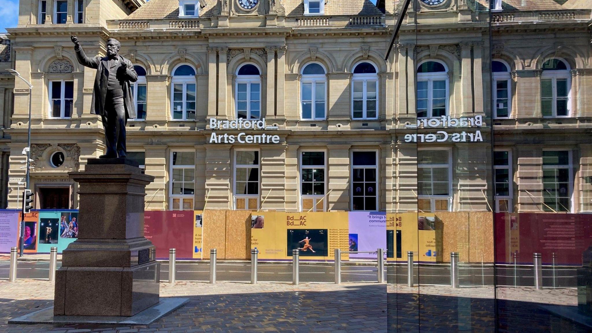 A Yorkshire stone building with multi-coloured hoardings covering its ground floor reflected in a glass window.