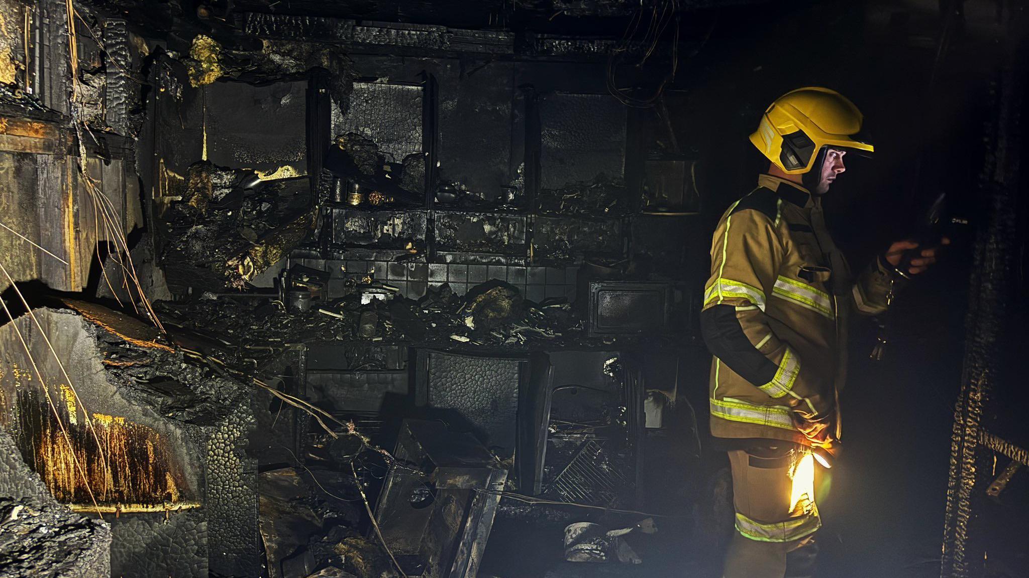 A firefighter with a yellow helmet is in the charred property, carrying a torch and holding equipment.