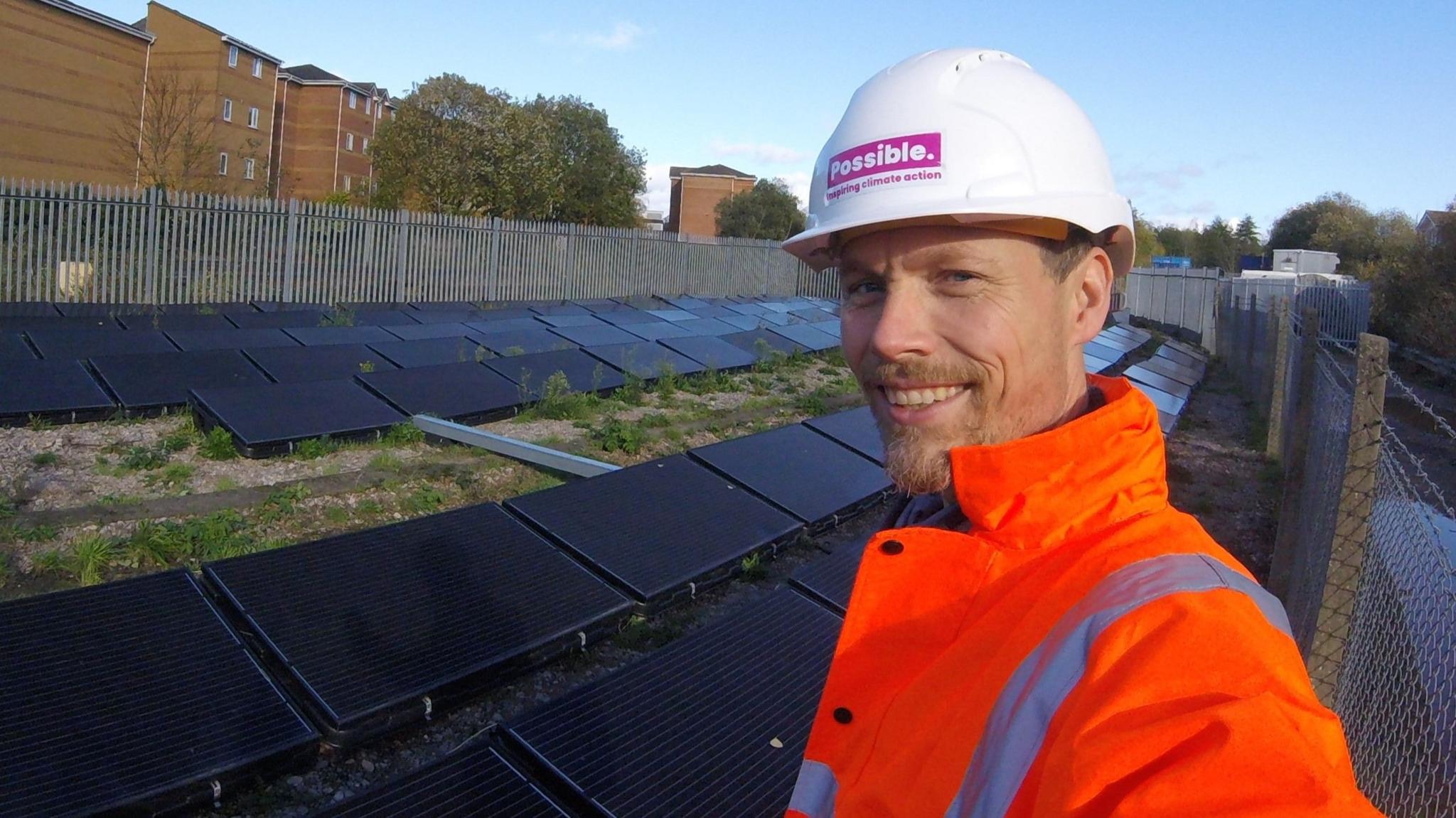 Leo Murray in a white helmet and orange hi-vis jacket takes a selfie with solar panels behind him.