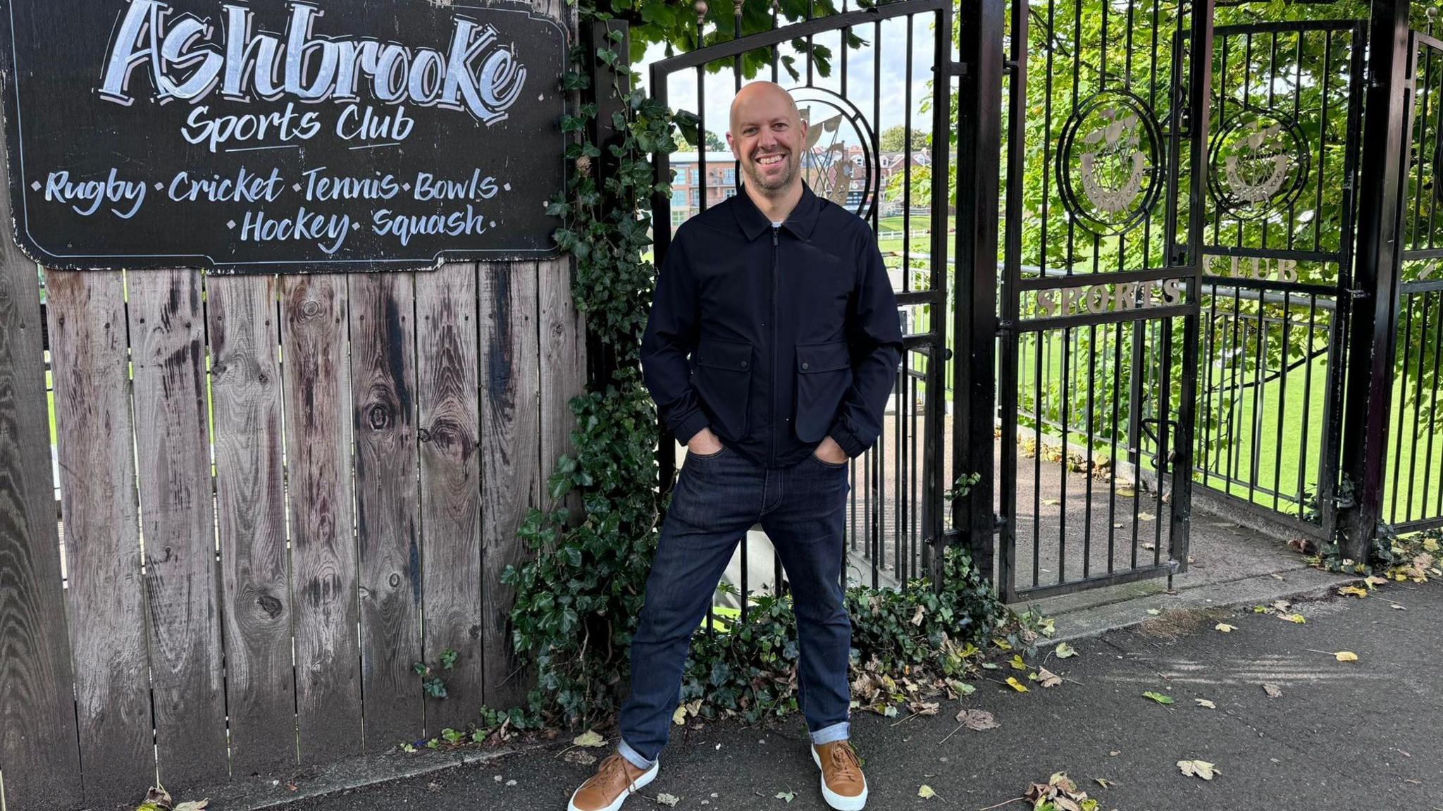 Ben Robinson, wearing a blue jacket and dark blue jeans, with a sign for Ashbrooke Sports club behind him. There are some railings and a wooden fence. 