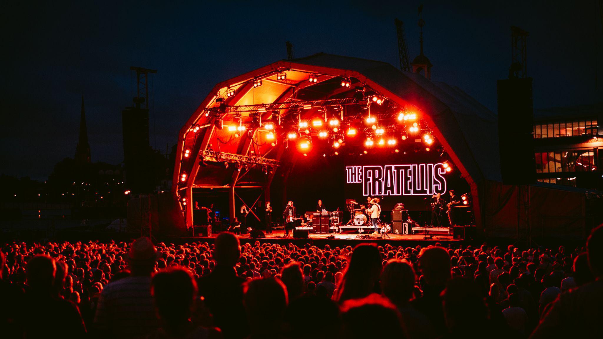 The Fratellis performing at Bristol Sounds. The stage is lit in red and the band is standing in front of their logo, projected on the back in capital letters. There is a huge crowd watching them.
