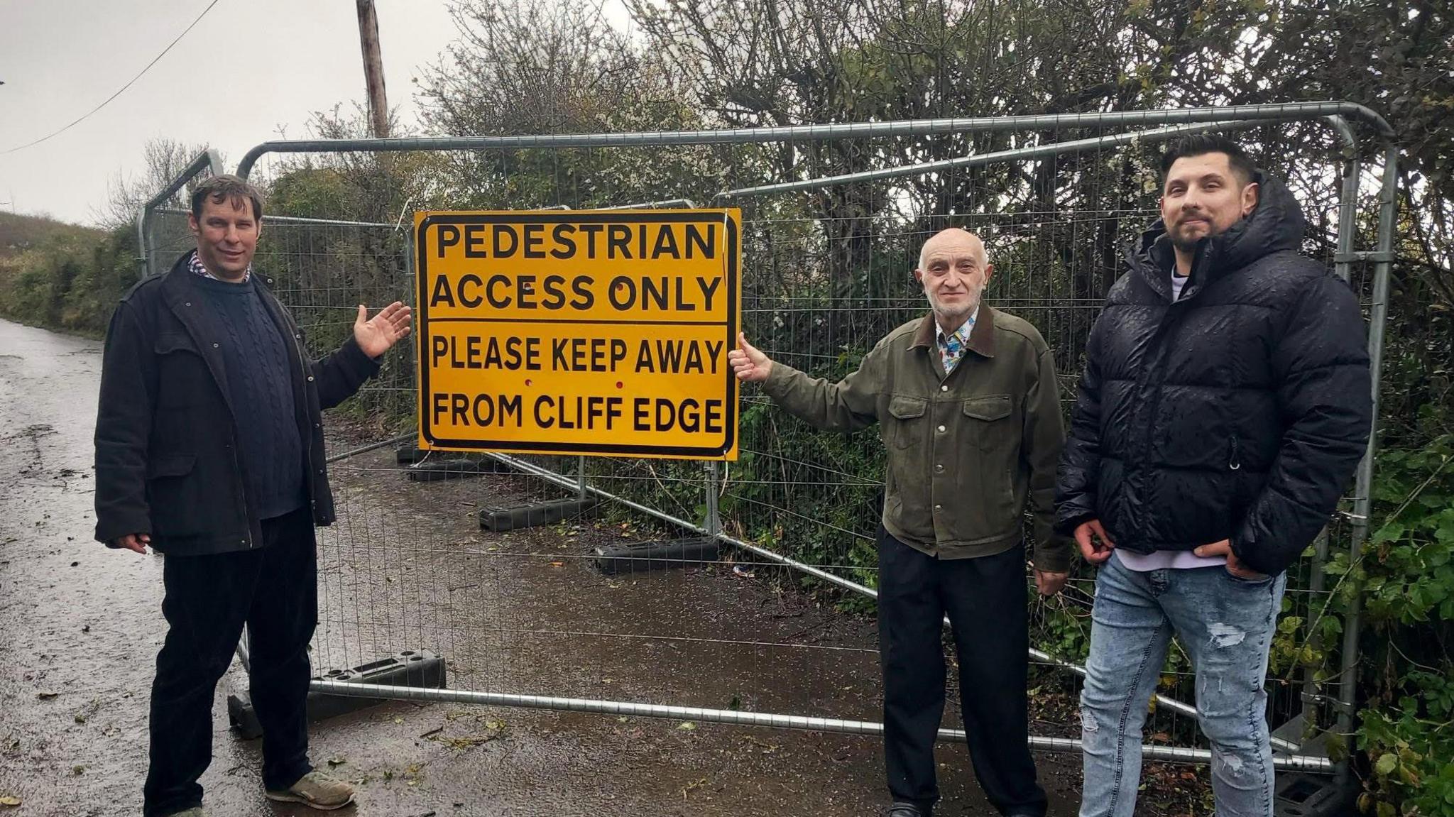 Three men stood on a cliff-top road. It has a big sign saying "Pedestrian Access Only - Please Keep Away from Cliff Edge"