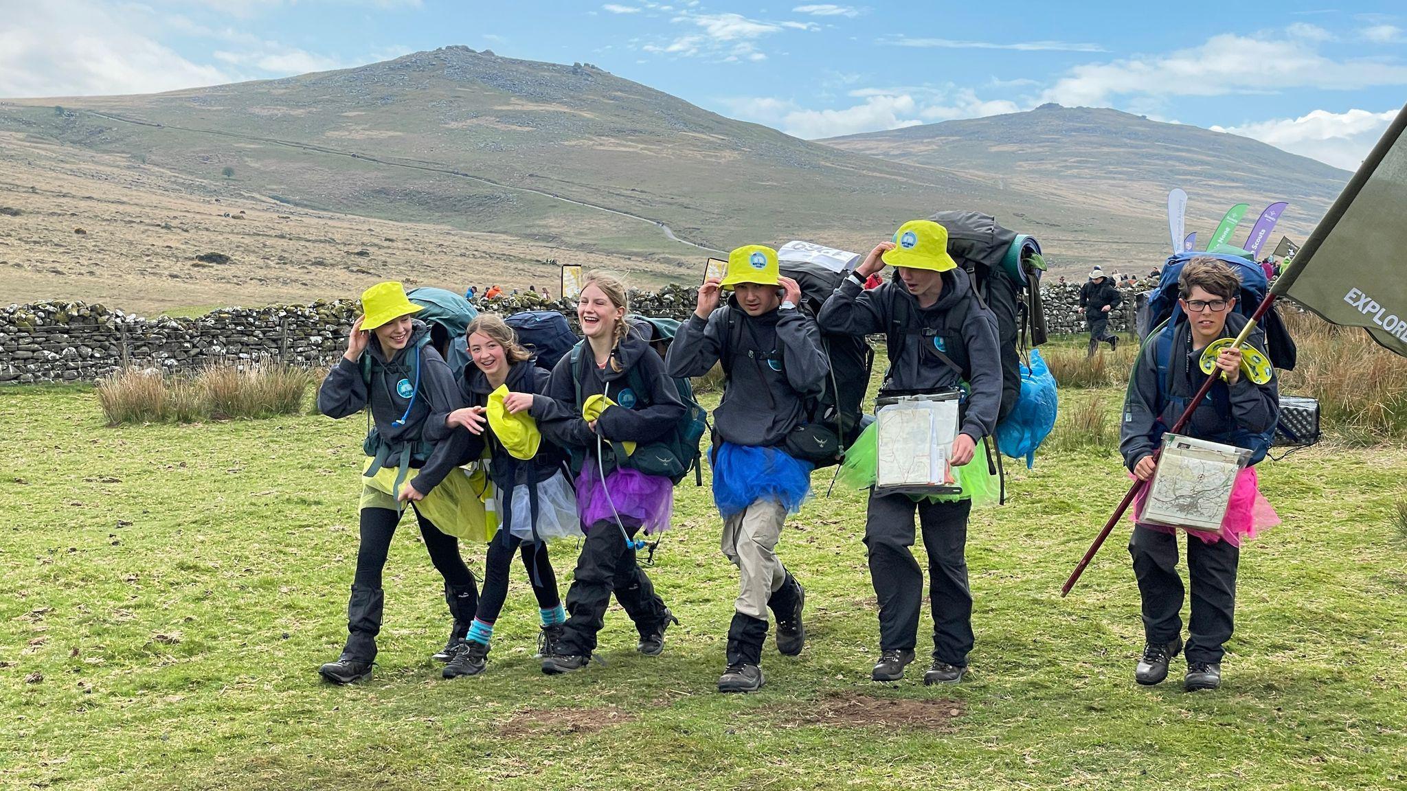 A group of six children is walking along a grass area with hills in the background. They are laughing and holding on to their yellow hats in the wind.