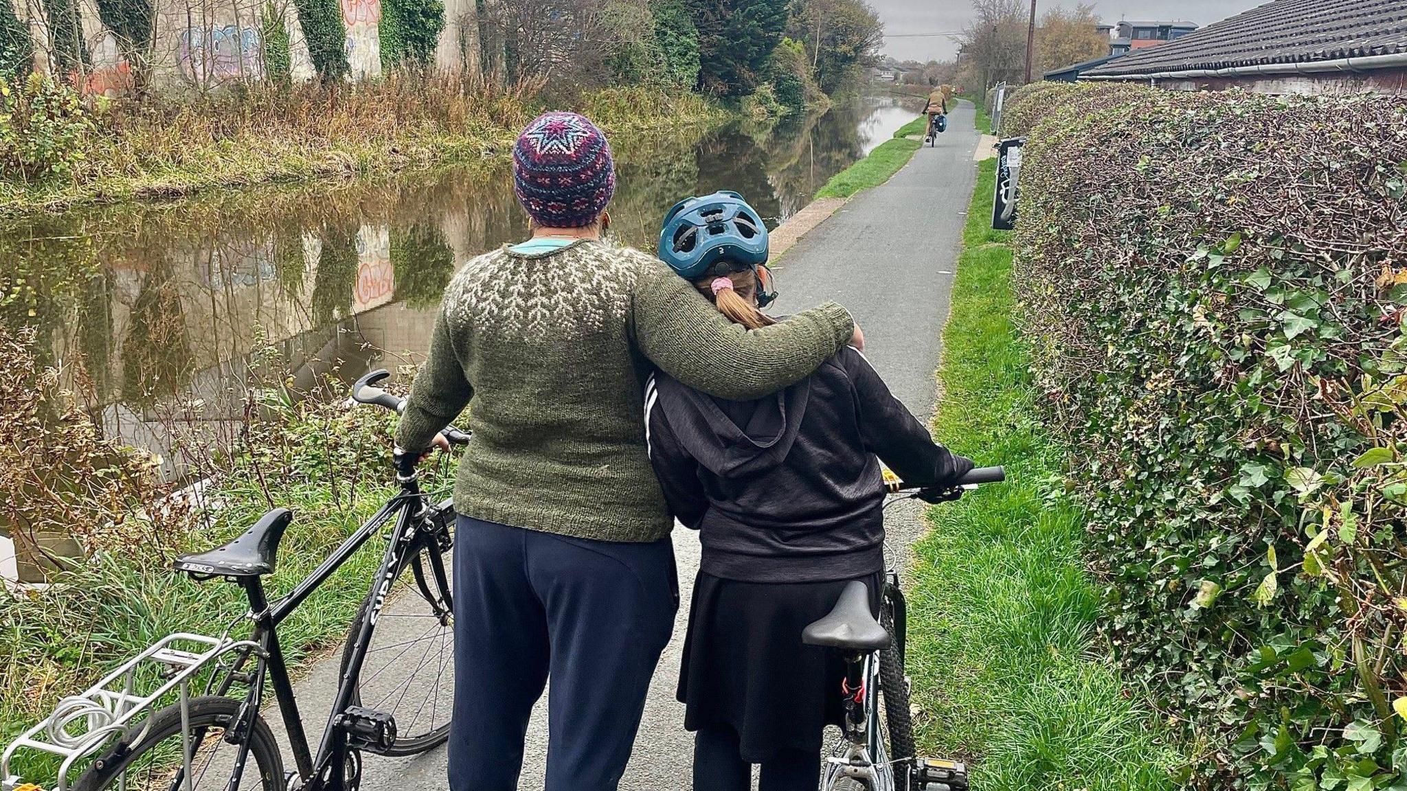 A mother stands with her back to the camera with her arm around her daughter's shoulders. They are standing next to the Union Canal and both are holding their bikes. The mum is on the left and is wearing a green knitted jumper and a colourful wooly hat and the daughter is wearing a cycling helmet and dark skirt and top.