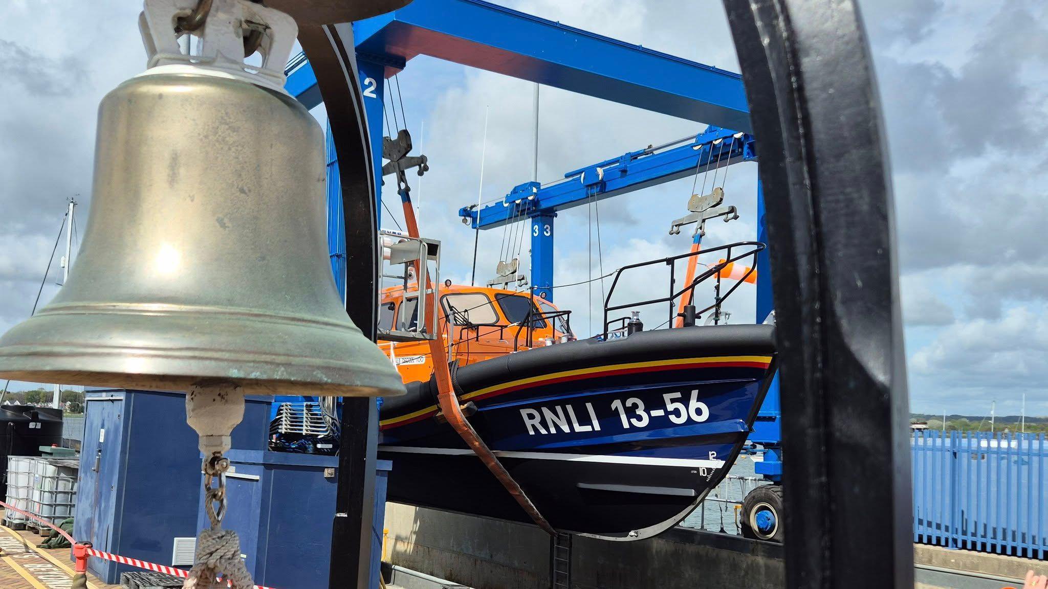 A large silver bell is in the top left-hand corner and behind it is an orange and blue RNLI lifeboat. It says RNLI 13-56 on it. It is suspended above the water on a crane. The sea can be seen in the distance.
