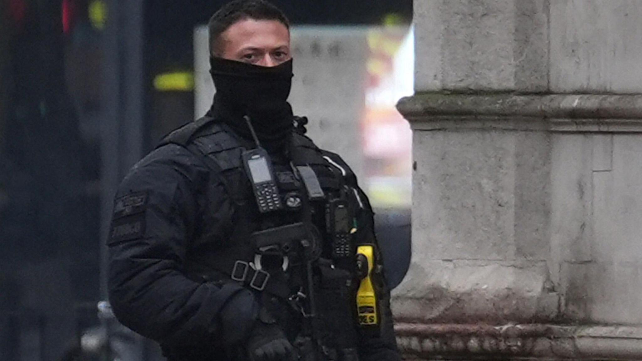 A male police officer, wearing black, face partly covered, holding a firearm.