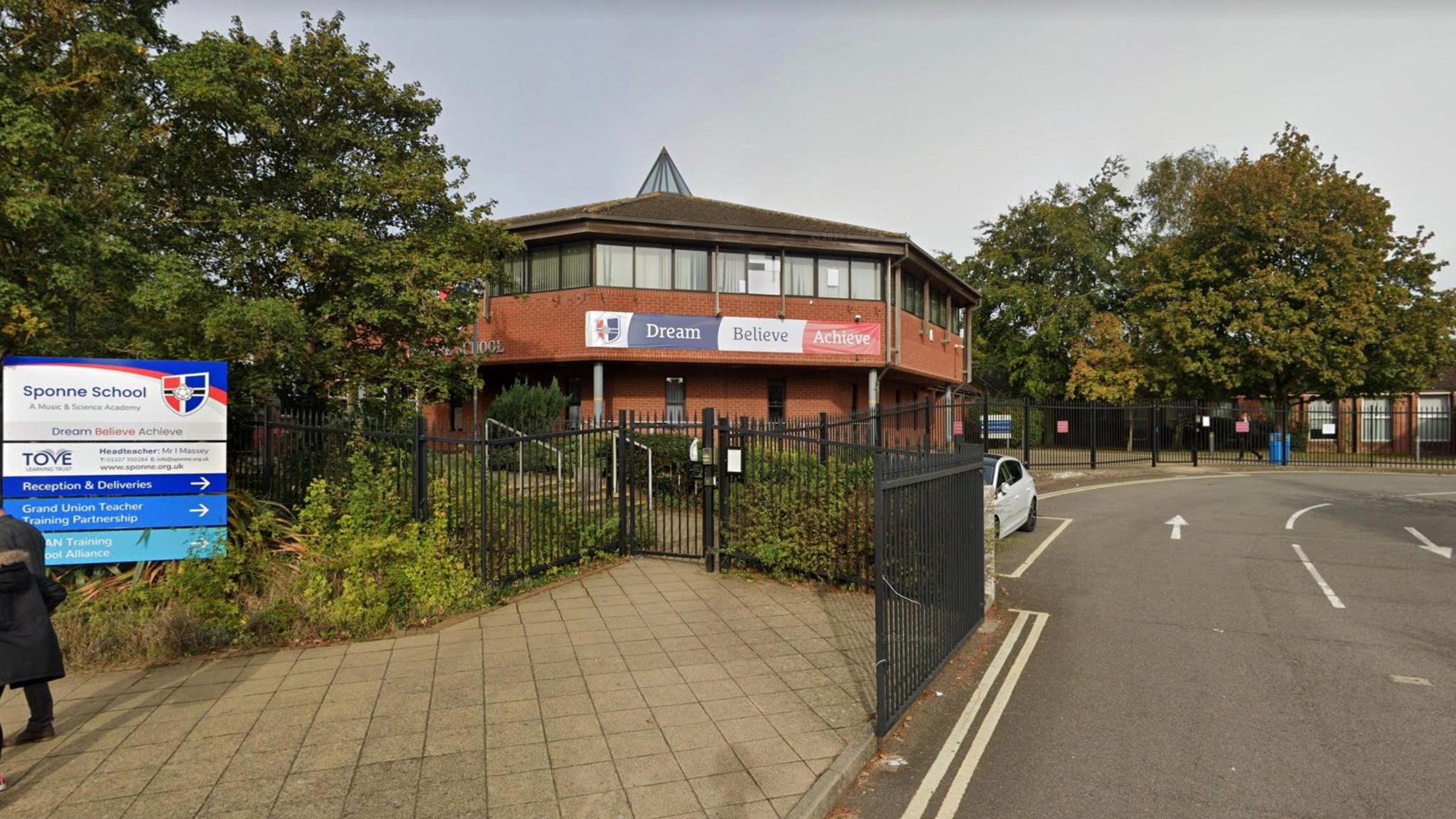 A picture of Sponne School in Towcester. A red brick angular building with windows and signage across the front. The building is shown in the middle of the image, with a paved path to the left and a sign showing "Sponne School". There is a road to the right of the picture