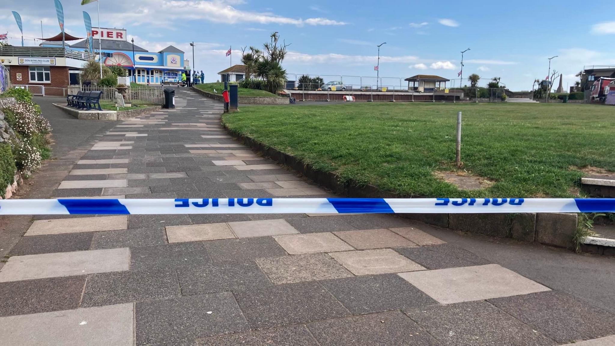 An image showing a path leading to a pier and some green space. There is police tape.