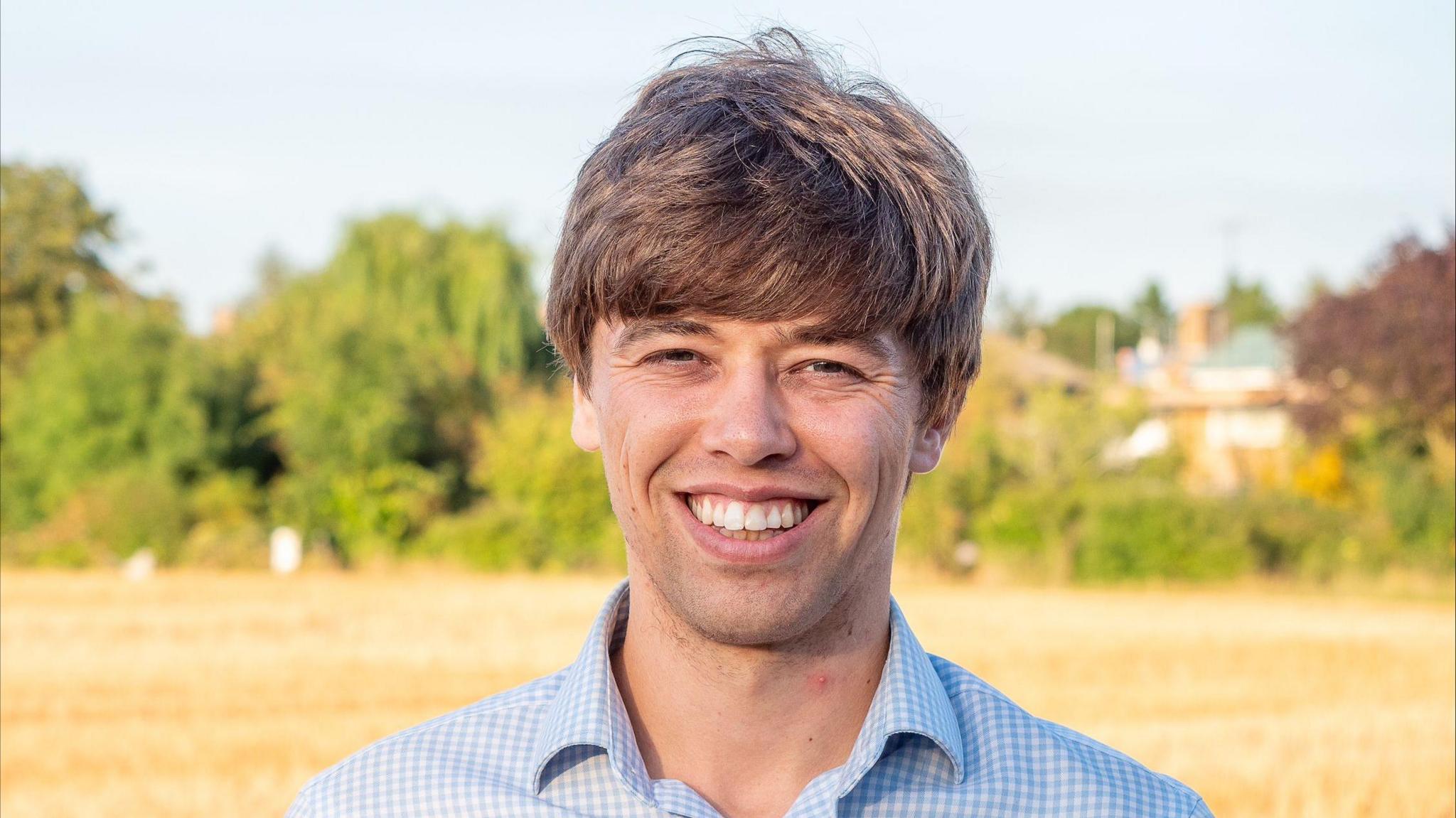 A smiling Cameron Holloway stares at the camera. He wears a blue and white checked shirt. In the background is a yellow field, green trees and a small building.