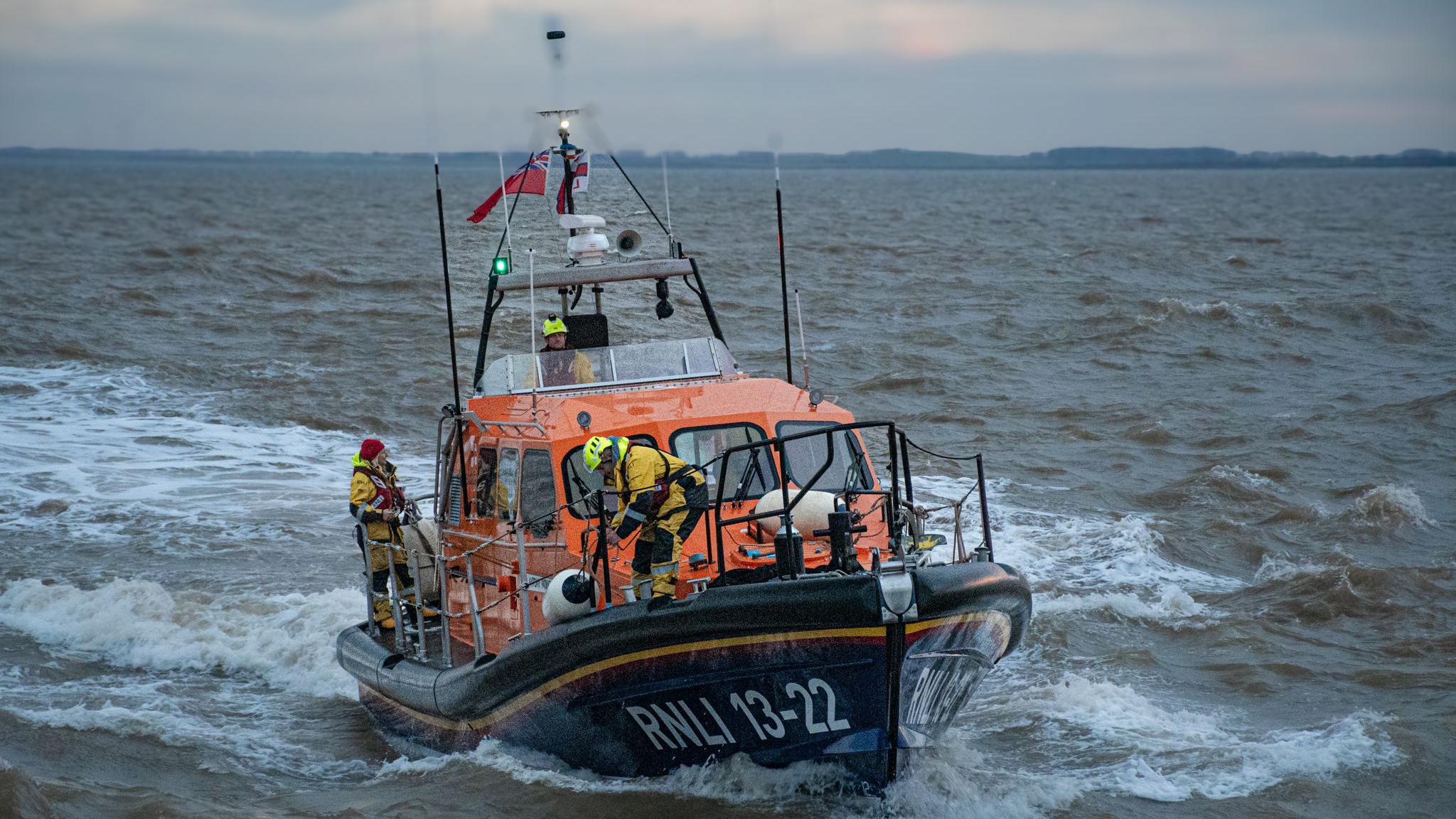 A lifeboat with three crew members in view, with one of them at the helm. "RNLI" is written on the side and the lifeboat is heading through water