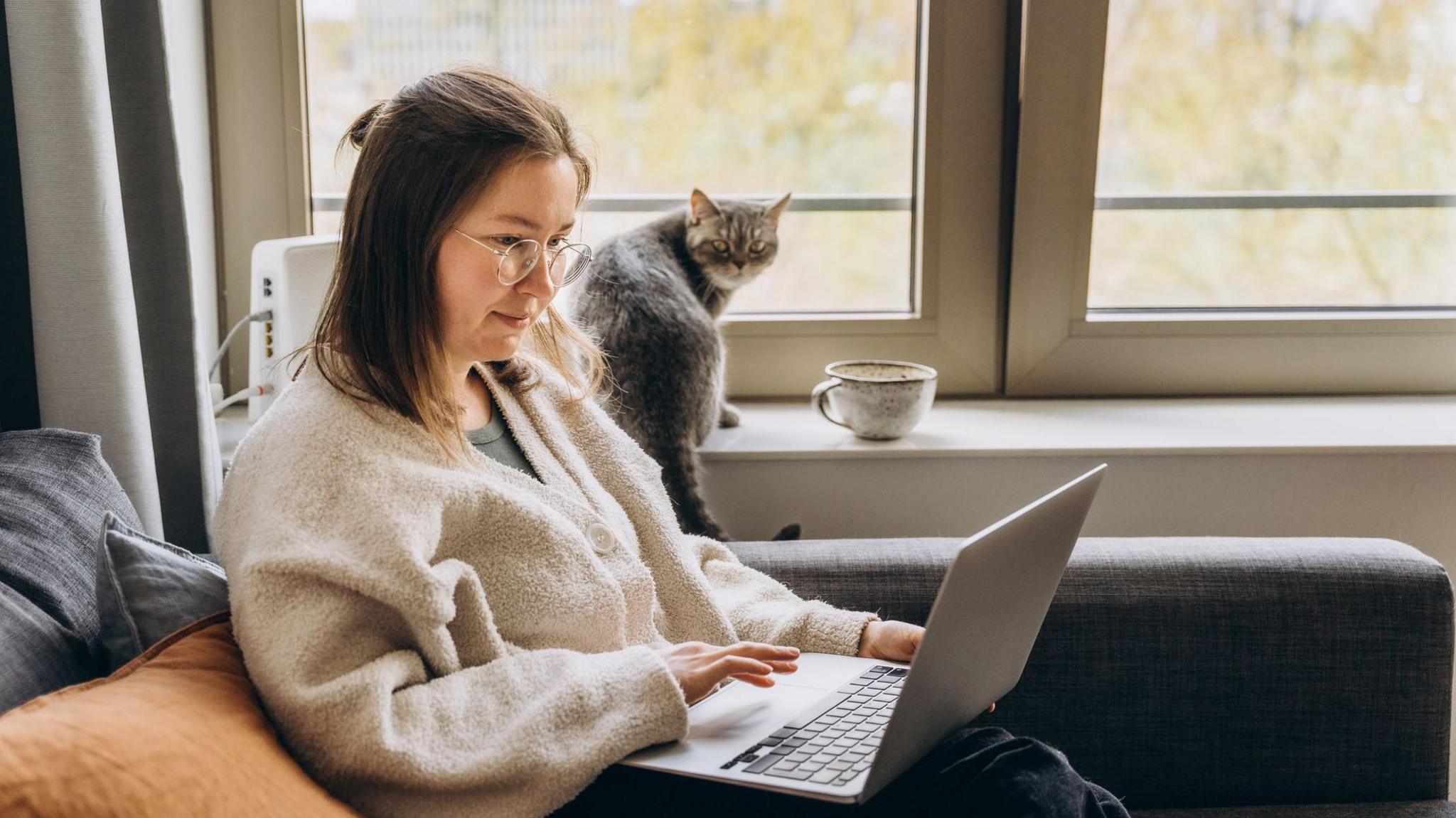 Woman sitting on a sofa using a laptop. A cat and a large ceramic mug are on the windowsill behind her. She has long brown hair, and is wearing glasses and a grey cardigan.