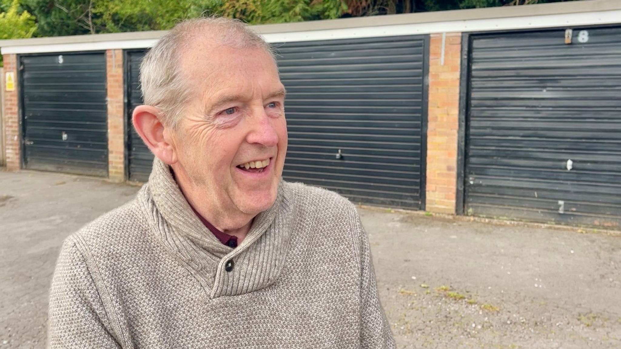 A man with short grey hair, who is wearing a beige woollen jumper and a maroon shirt, is standing in front of four black garages.
