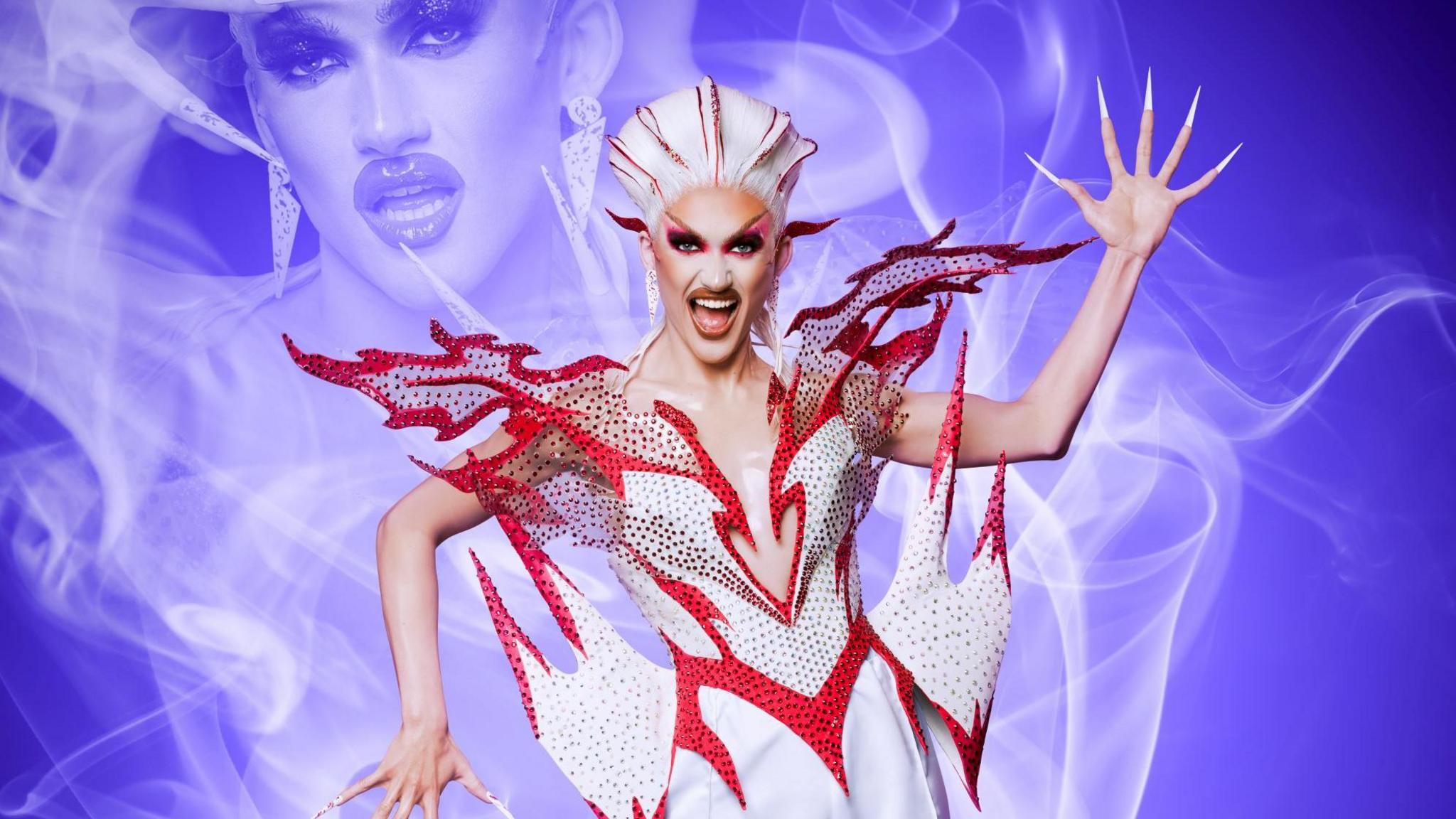 Kyran Thrax wearing a red and white outfit. The outfit has a red border which look like flames coming off the outfit. They have long nails and dramatic makeup. 