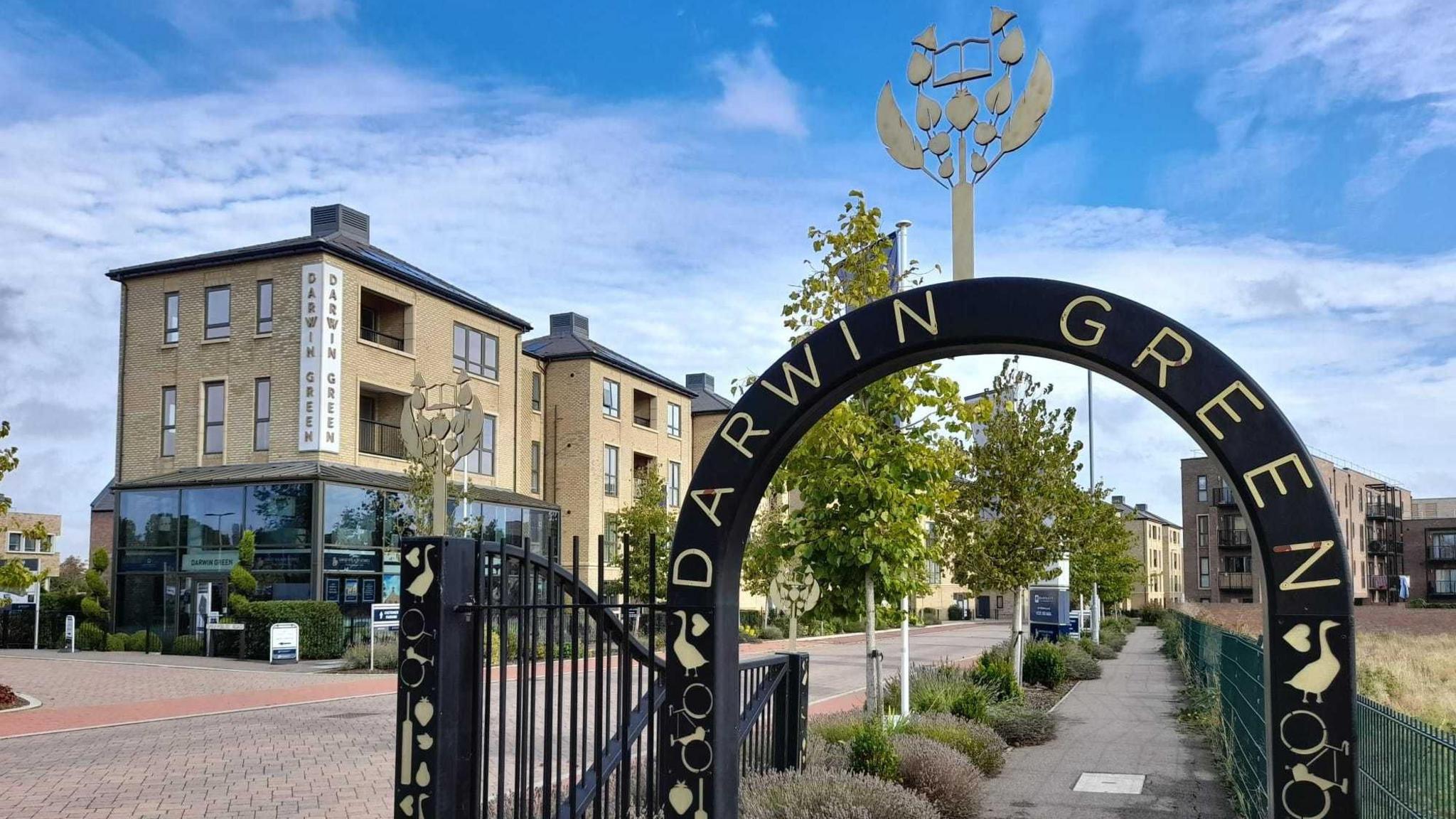 A Crescent shaped entrance gate with the words Darwin Green on it, is over a path. Beyond that are newly built 4 storey flats, trees and bushes.   