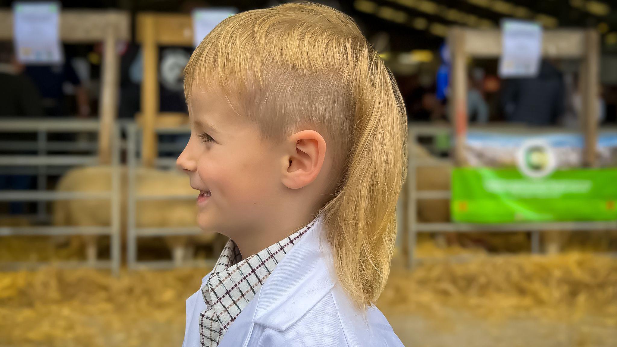 Boy in a sheep shed wearing a white coat for showing and with a long blonde mullet