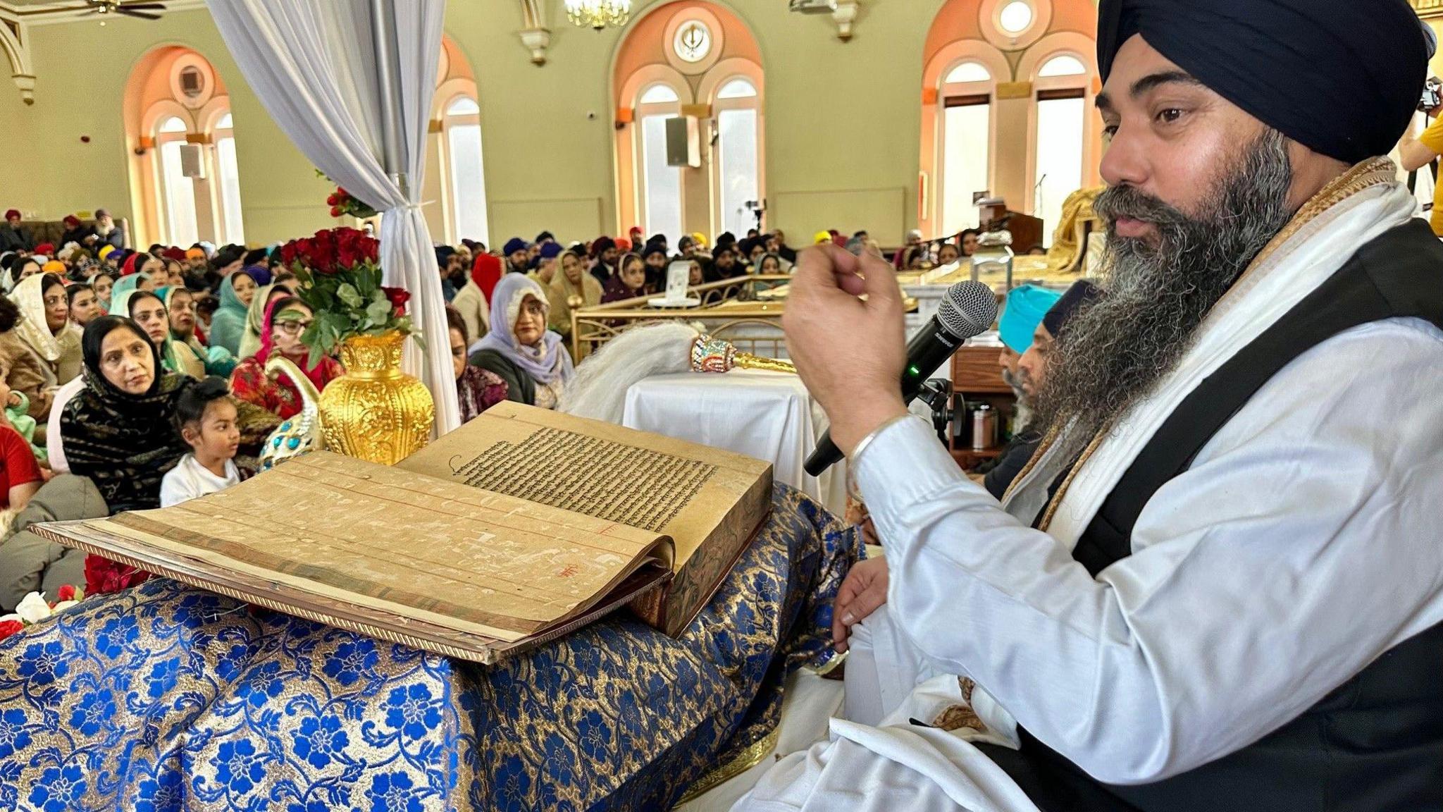 The priest is reading into a microphone. The  Guru Granth Sahib is in front of him. The crowd is sitting on the floor facing him.