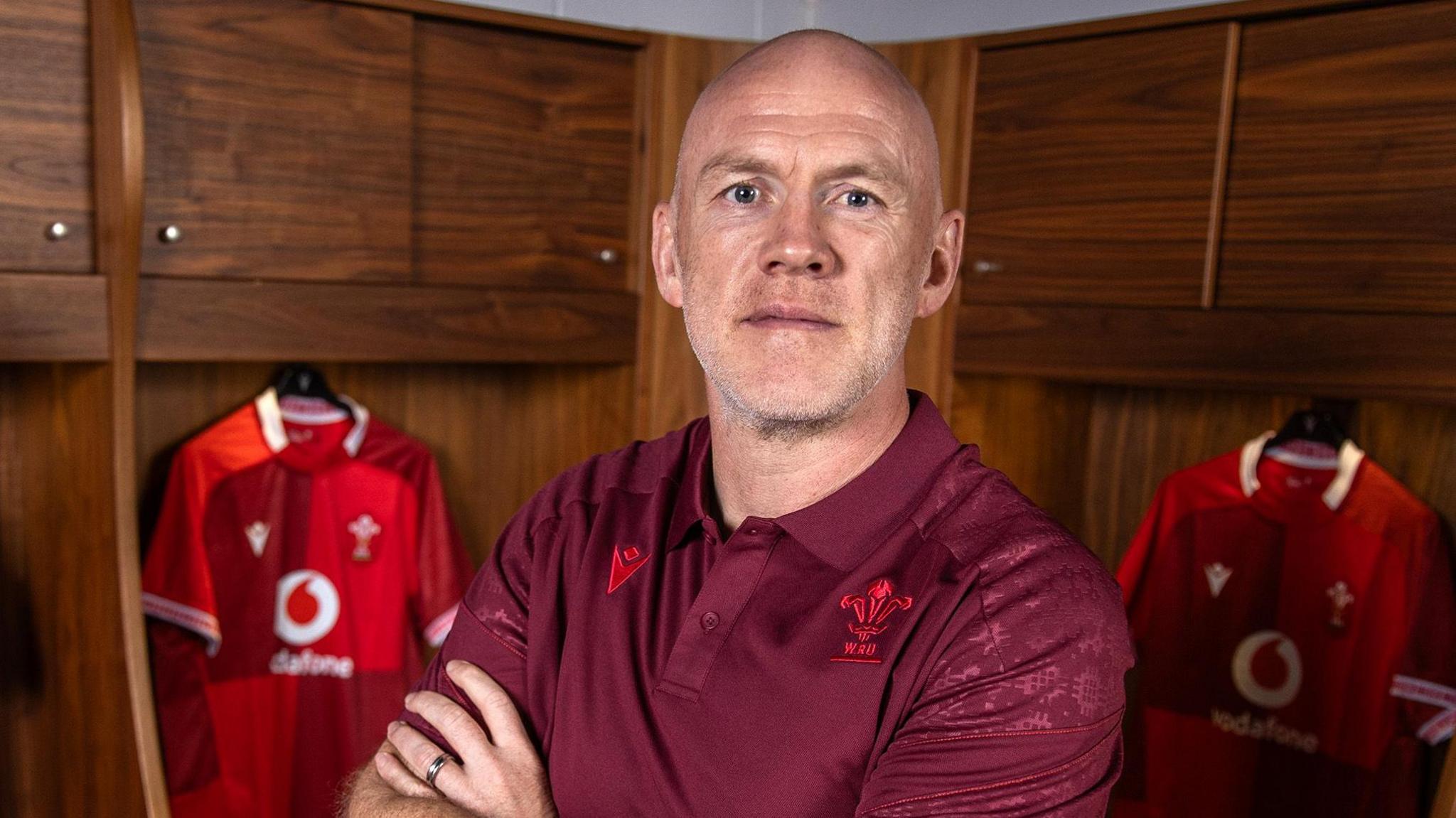 Steve Tandy posing with his arms folded in front of Wales jerseys in locker room