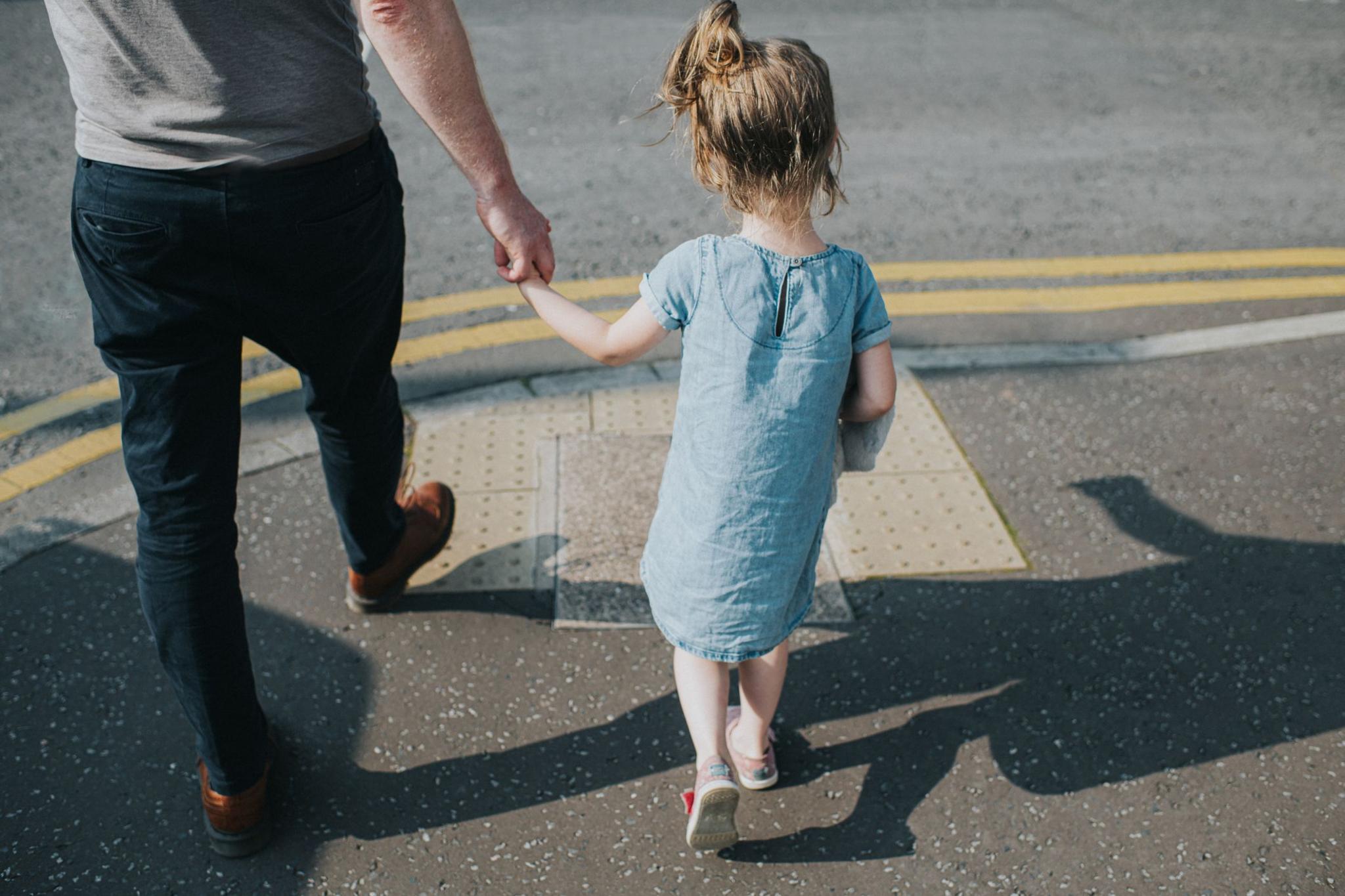 A man wearing brown shoes and dark trousers and a young girl wearing a light blue dress walk hand in hand on a pavement casting shadows on the ground as they come to a road