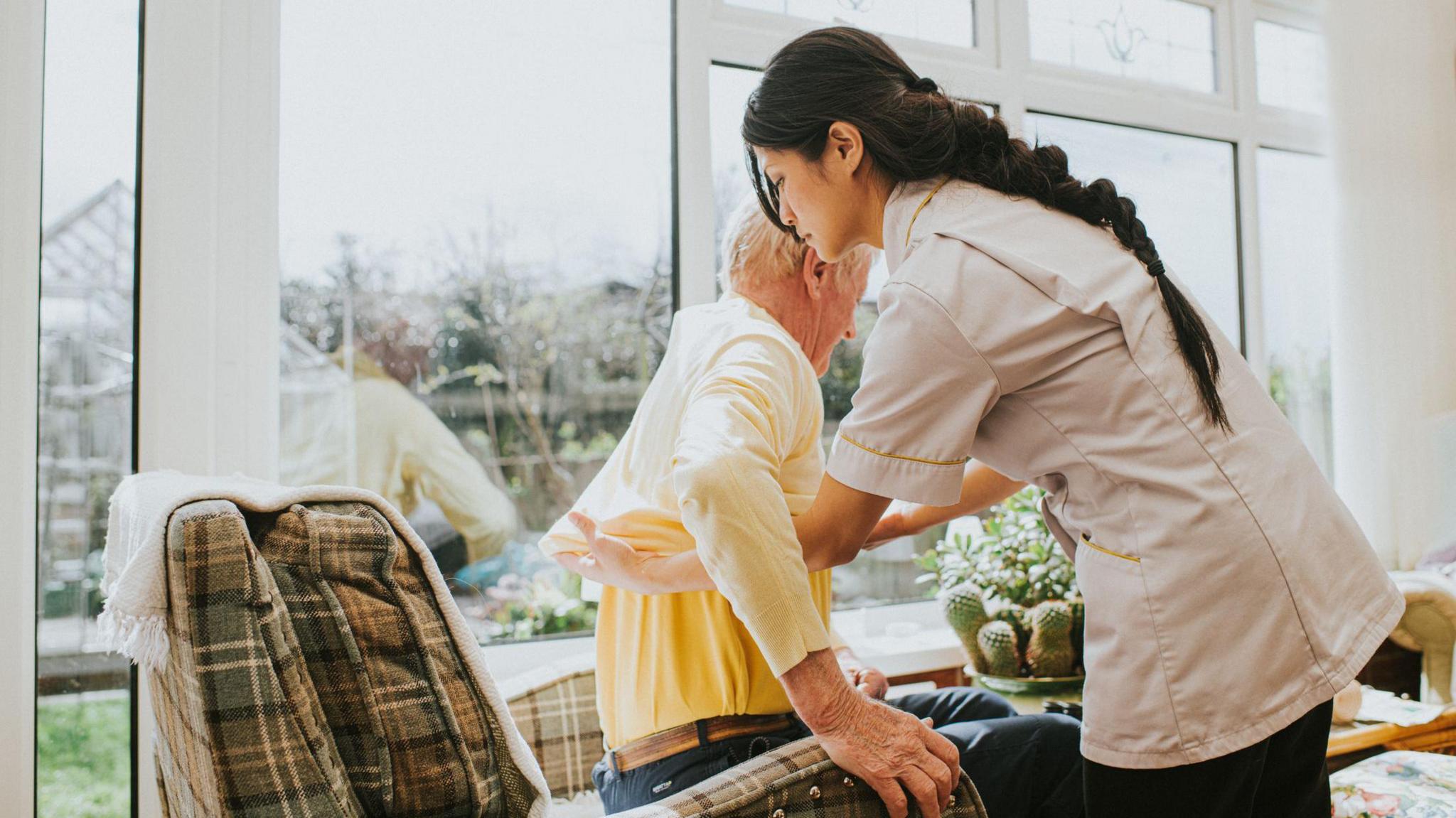 a care worker wearing a medical tunic helps and elderly man into a chair in his home