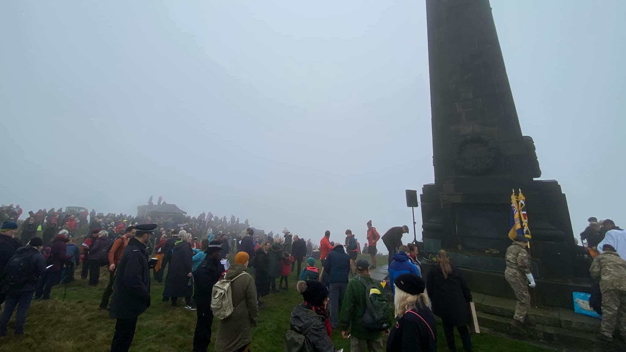 Hundreds of people gather at the base of the monument in fog. Military personnel in camouflage uniform place ceremonial standards with the red, white and blue union flag on the monument base.