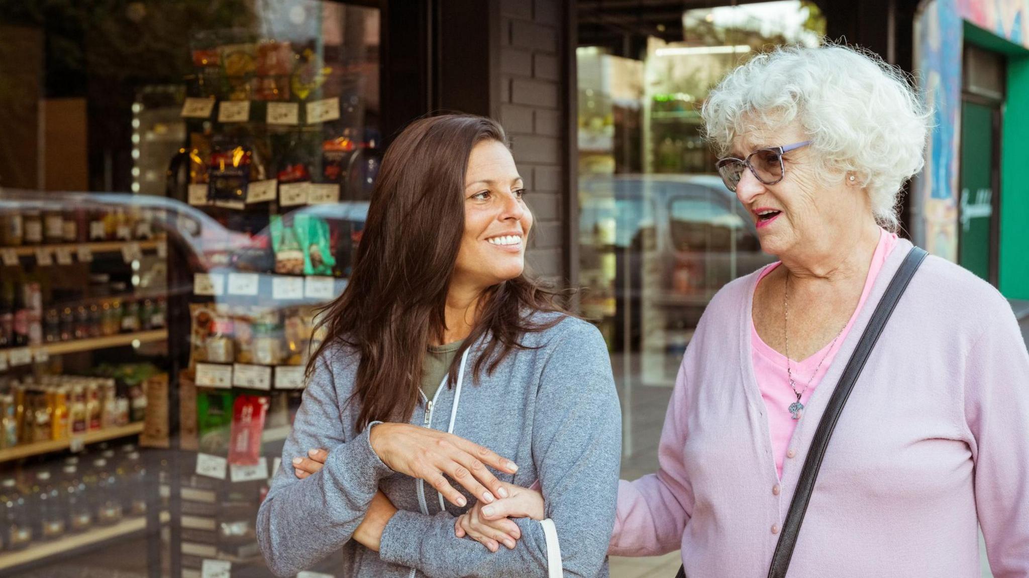 Smiling senior woman going to shopping with female, walking footpath arm in arm in the town.