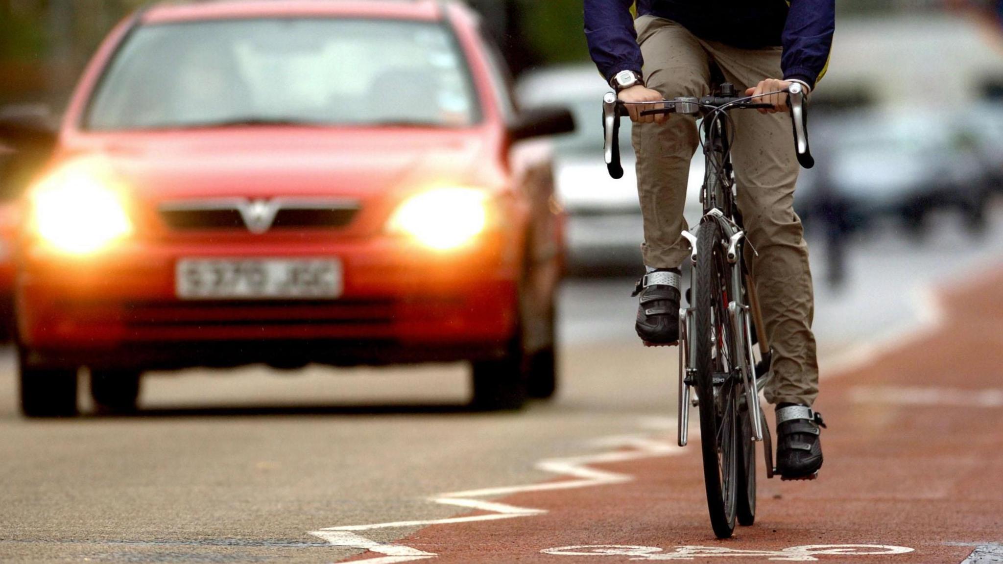 A cyclist rides on a red cycle land which is next to a road. A red car, with it's headlights on is to the left.