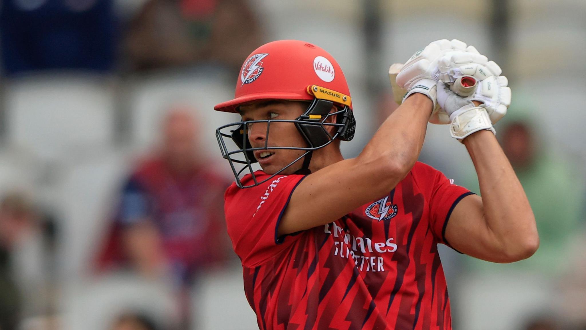 Chris Green wearing Lancashire's red T20 kit, plays a big shot while batting with his gloves and helmet on